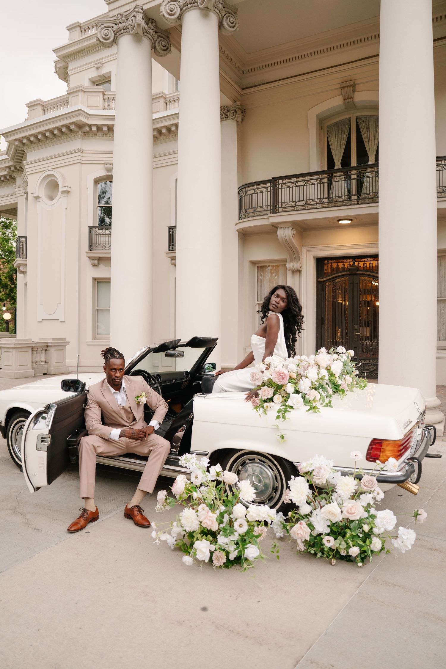 Elegant wedding couple posing with a white vintage convertible adorned with pink floral arrangements outside a grand mansion.