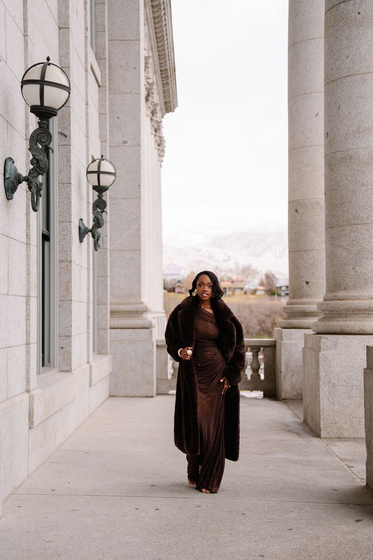 Elegant woman in brown velvet gown and fur coat walking between stone columns of a grand building.