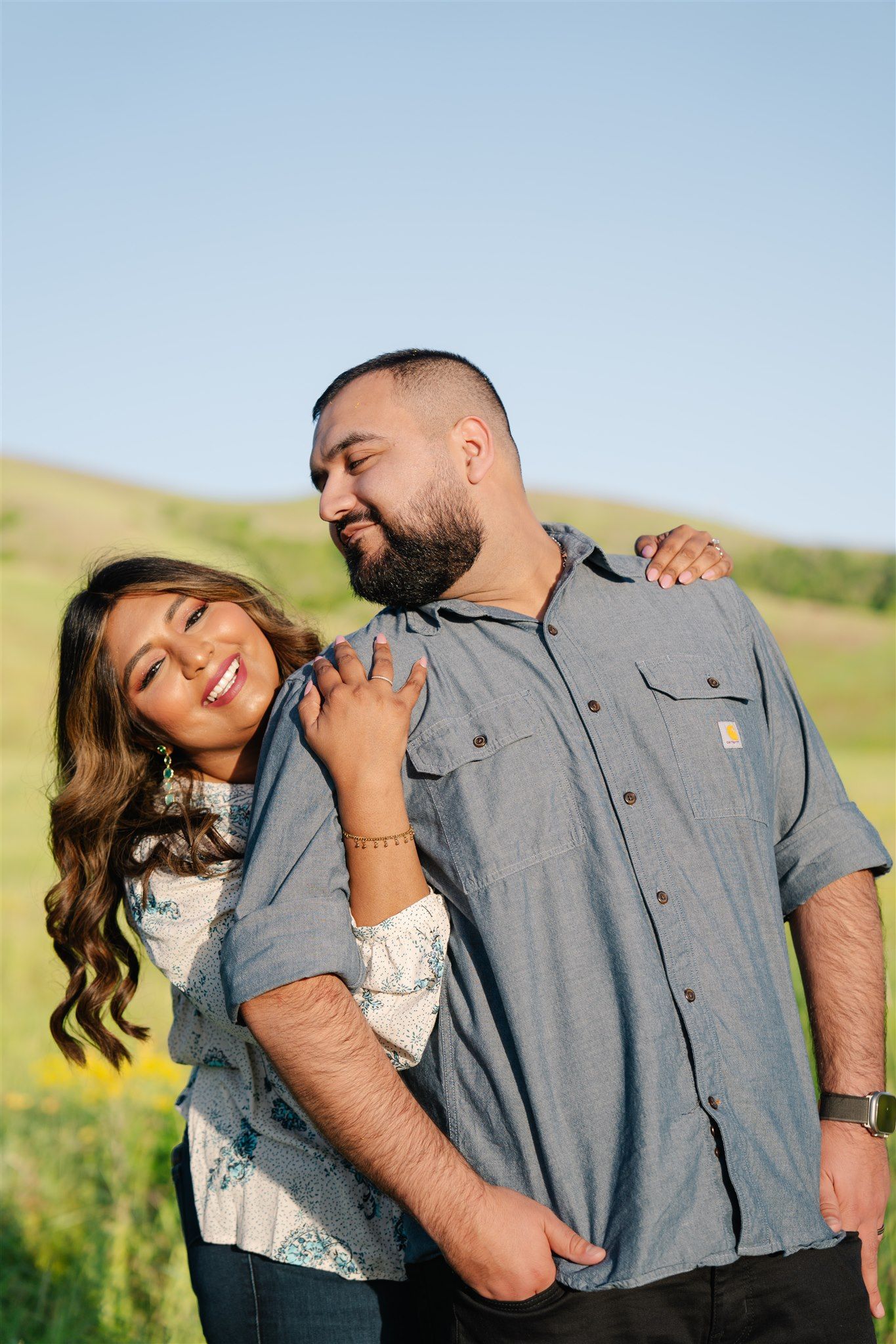 Playful couple laughing and posing in outdoor field with wildflowers and blue sky.