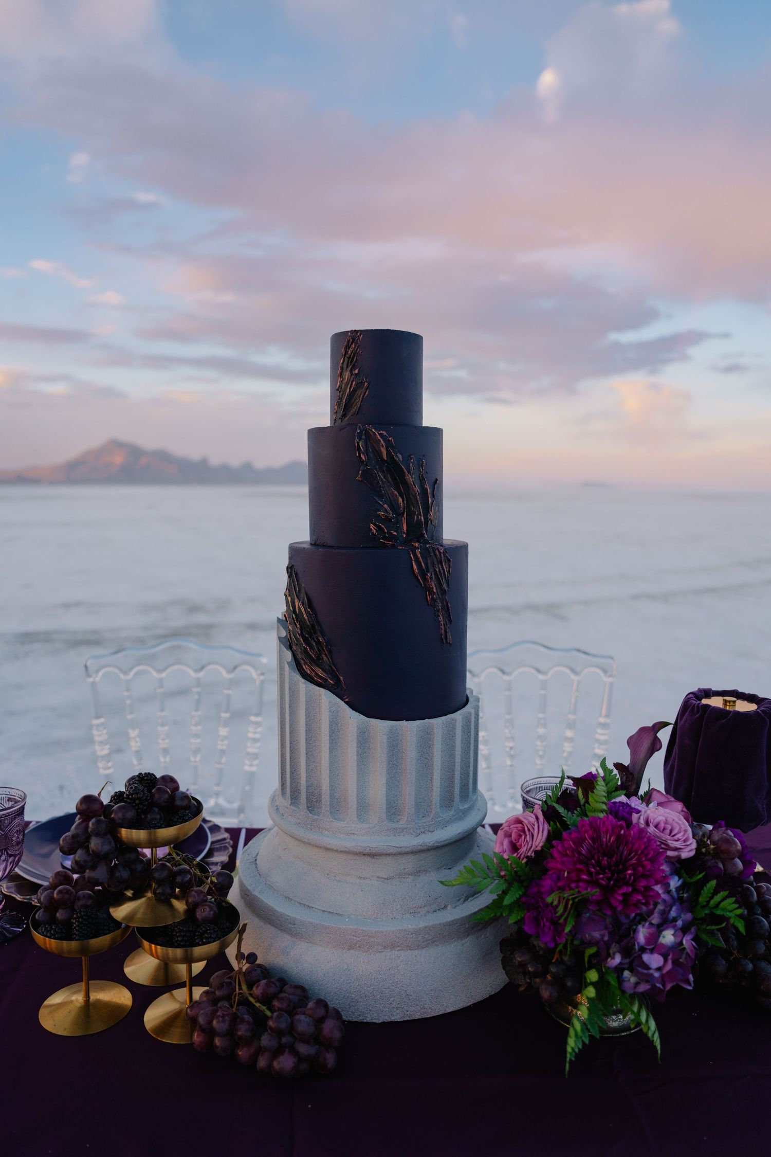 Elegant five-tier black and silver wedding cake with gold details on a pedestal, displayed outdoors at sunset by the ocean.