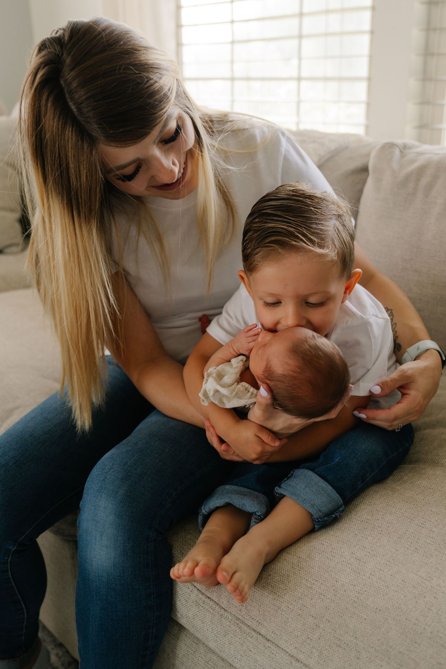 A young mother sits on a couch smiling as her toddler son gently kisses their newborn baby sibling.