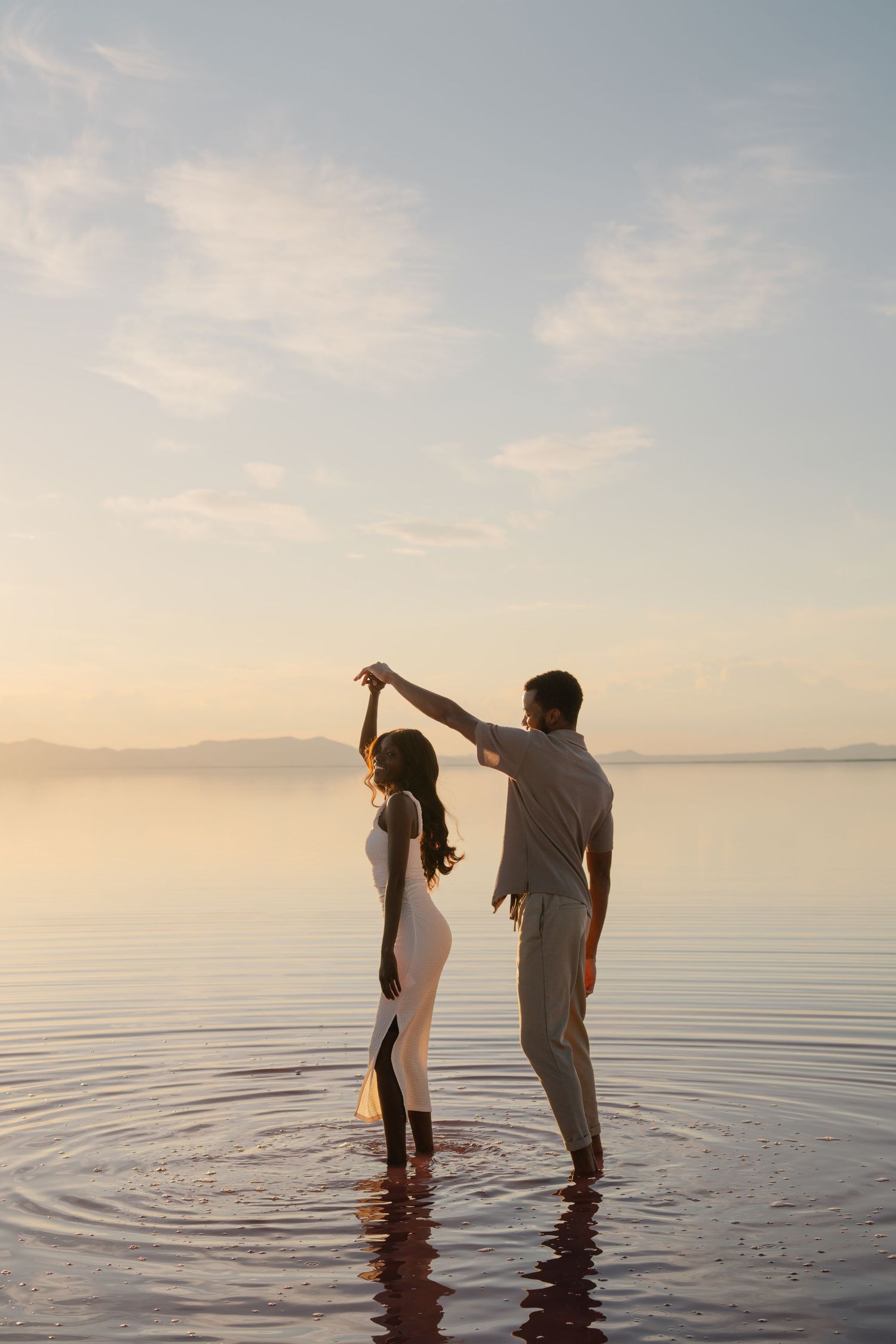Two people in white clothing dance and create ripples in shallow water at dusk.