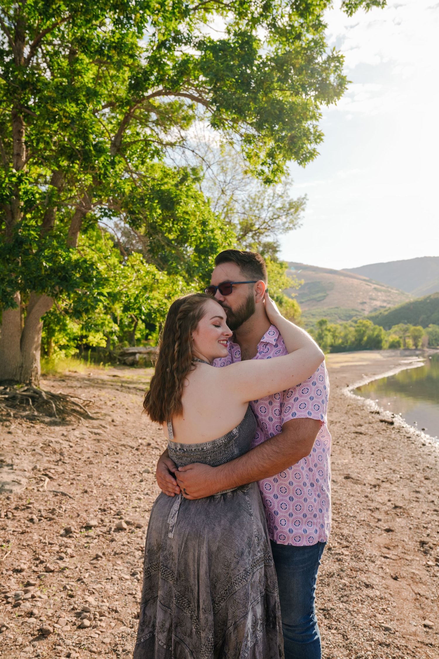 A romantic embrace by a lake with mountains and trees in the background.