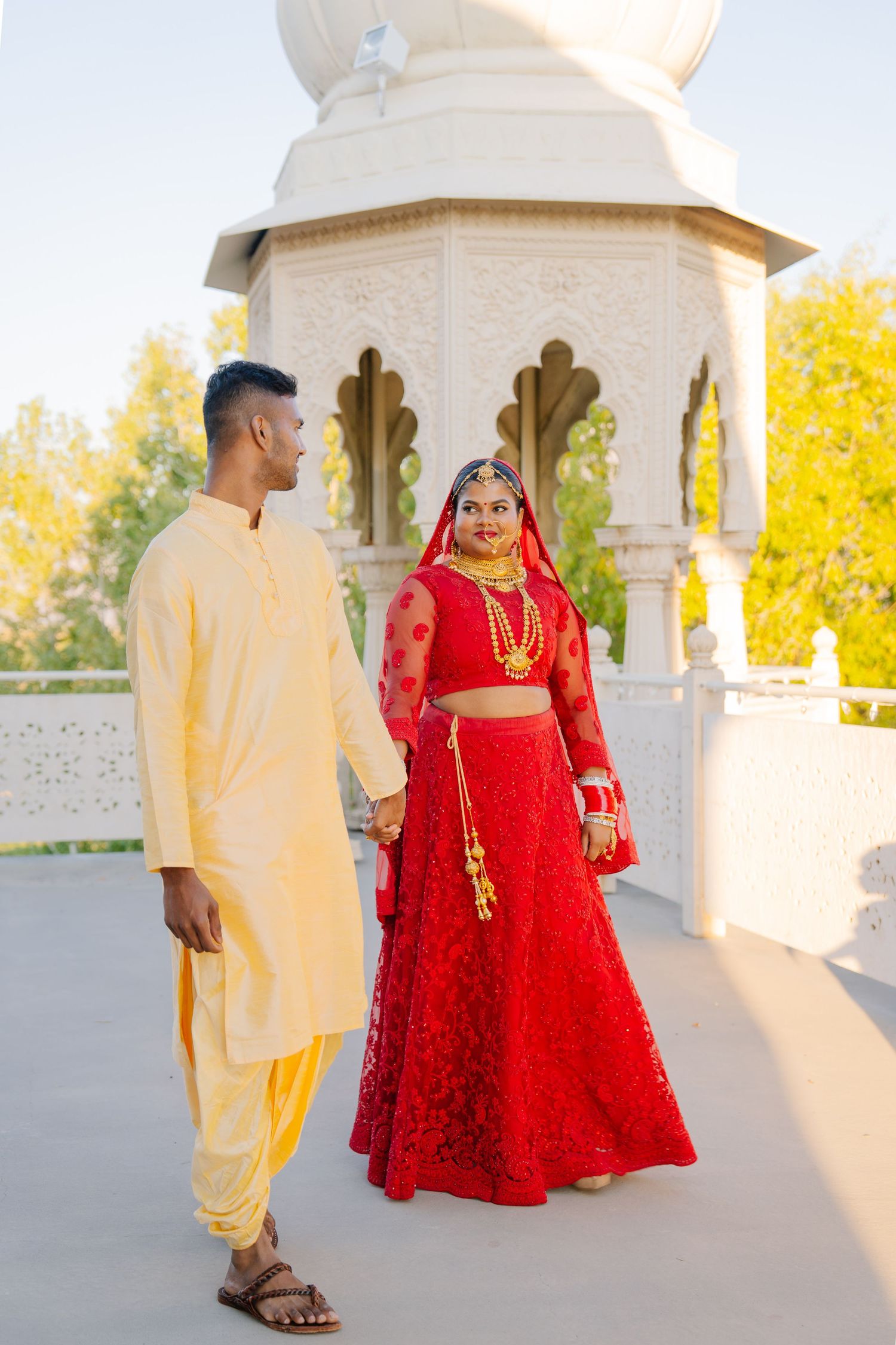 A couple in traditional Indian wedding attire walks through a marble pavilion.