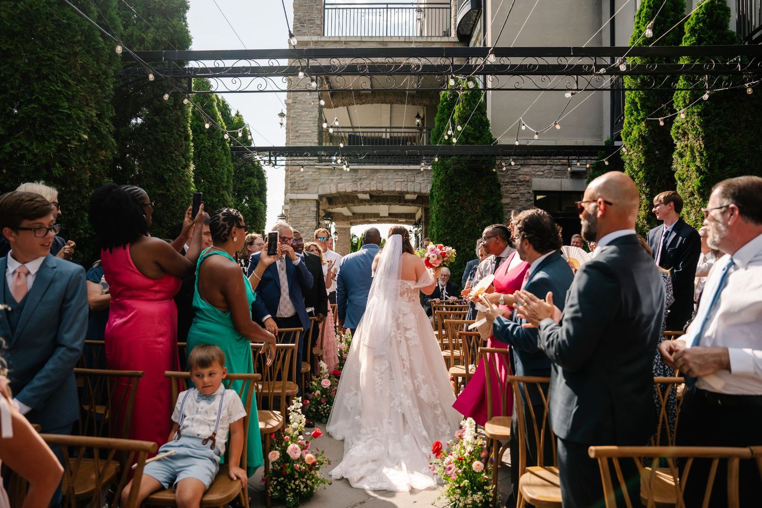 Wedding guests line the aisle as newlyweds exit their outdoor ceremony under floral decorations.