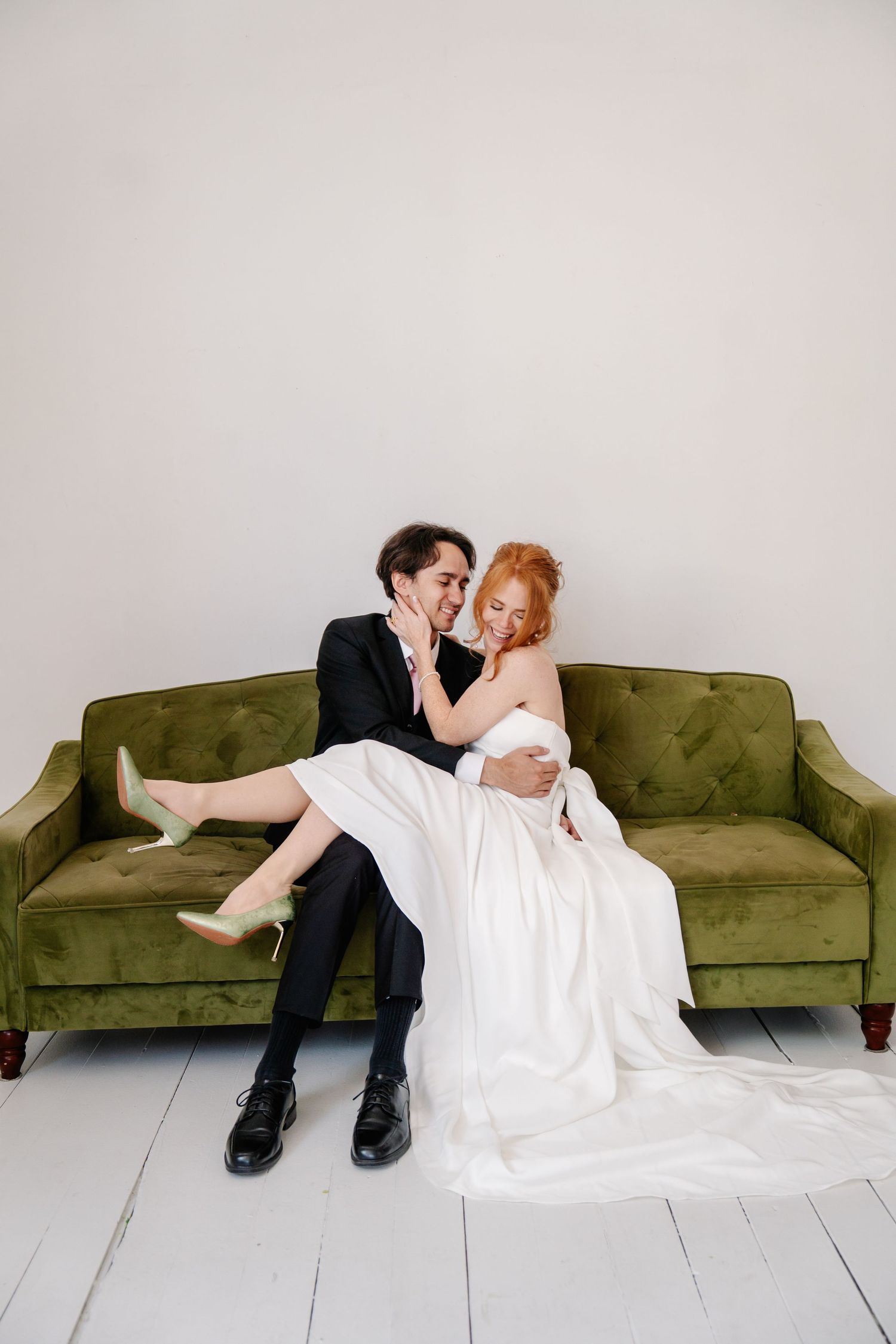 A couple lounges playfully on a green velvet couch in a minimalist setting.