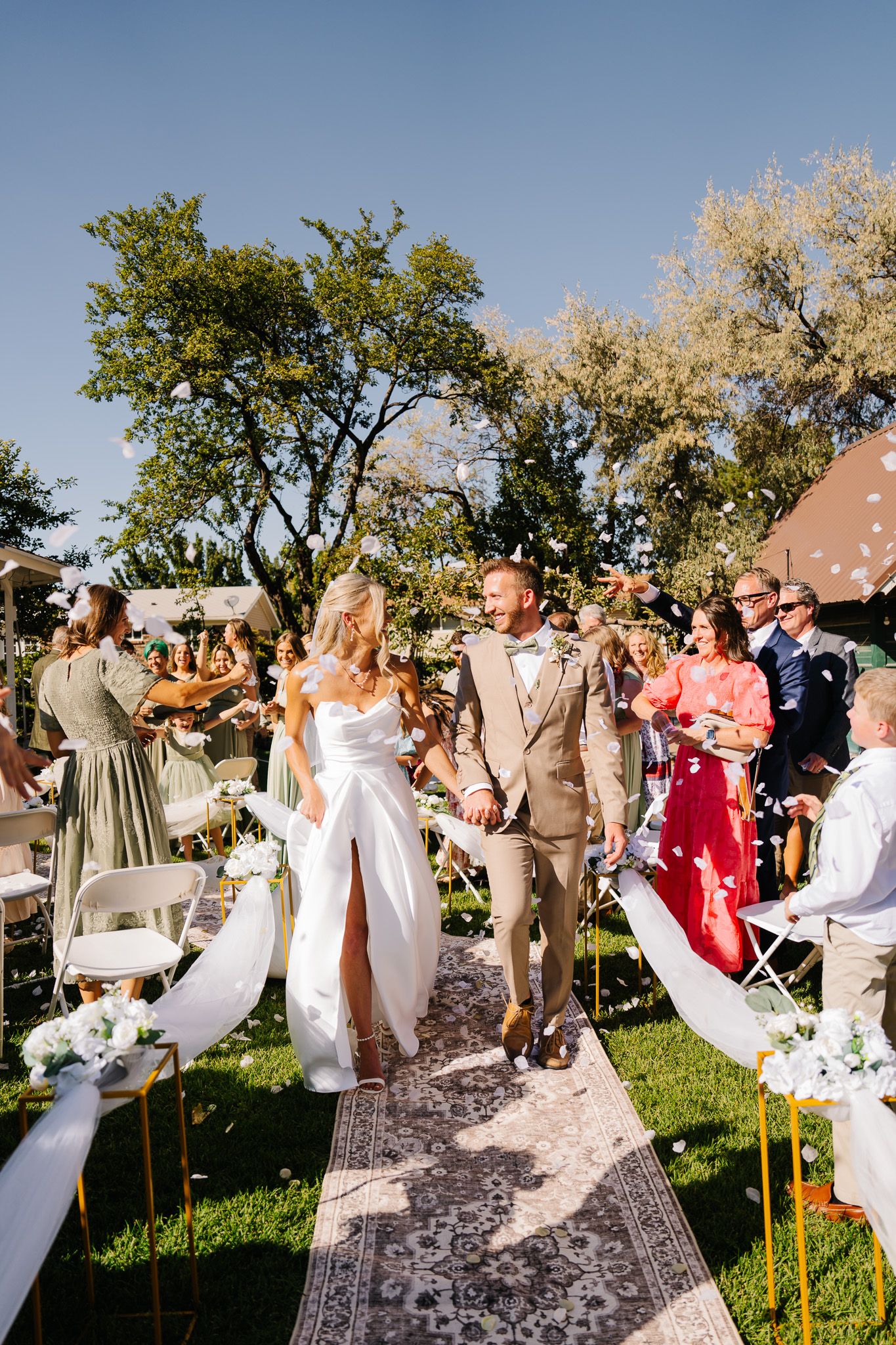 Wedding party celebrates joyfully on outdoor lawn with trees in background.