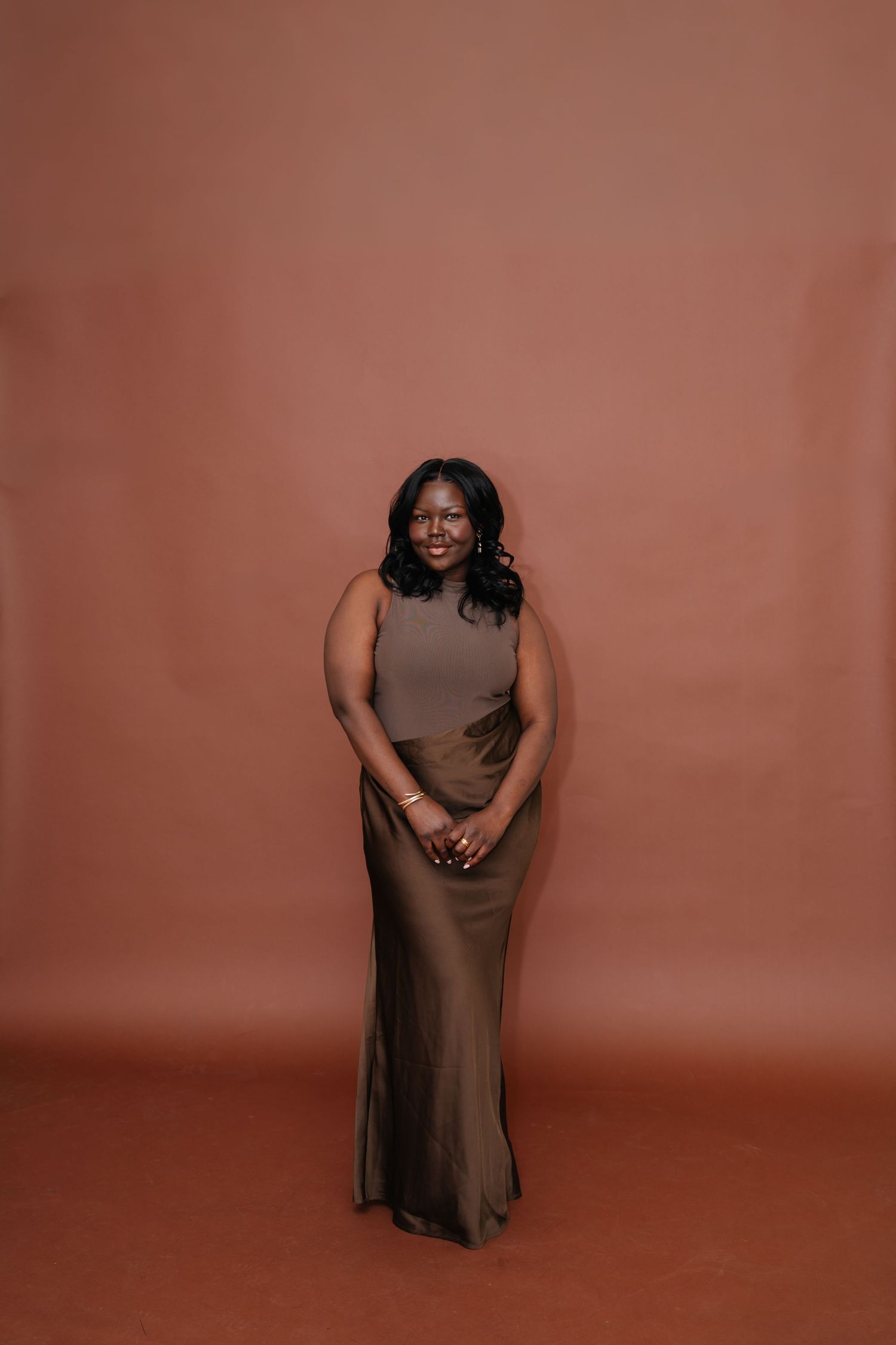 Woman in olive sleeveless top and maxi skirt smiling against a warm terracotta studio background.