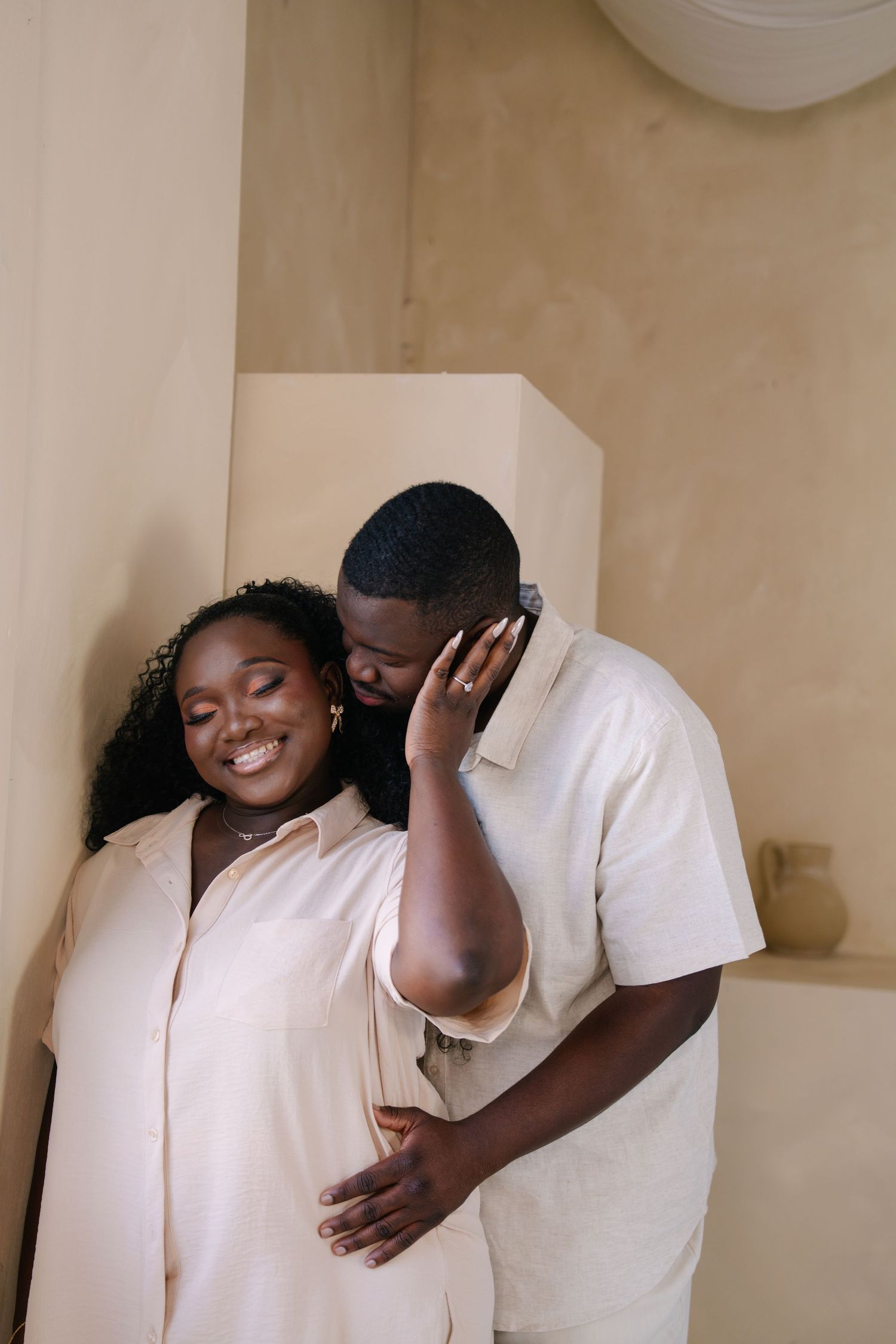A couple sharing a tender moment, the man gently kissing the woman's temple while she smiles, both in neutral tones.