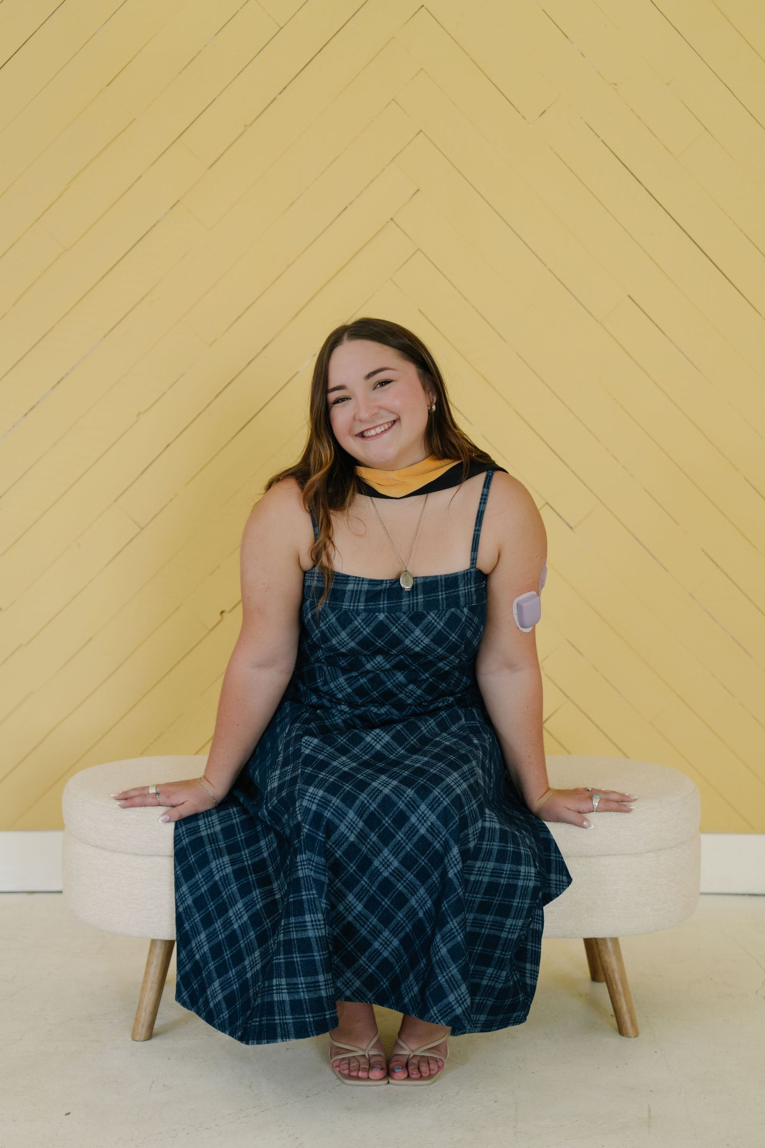 Smiling young woman in plaid dress seated on ottoman against yellow chevron wall backdrop.