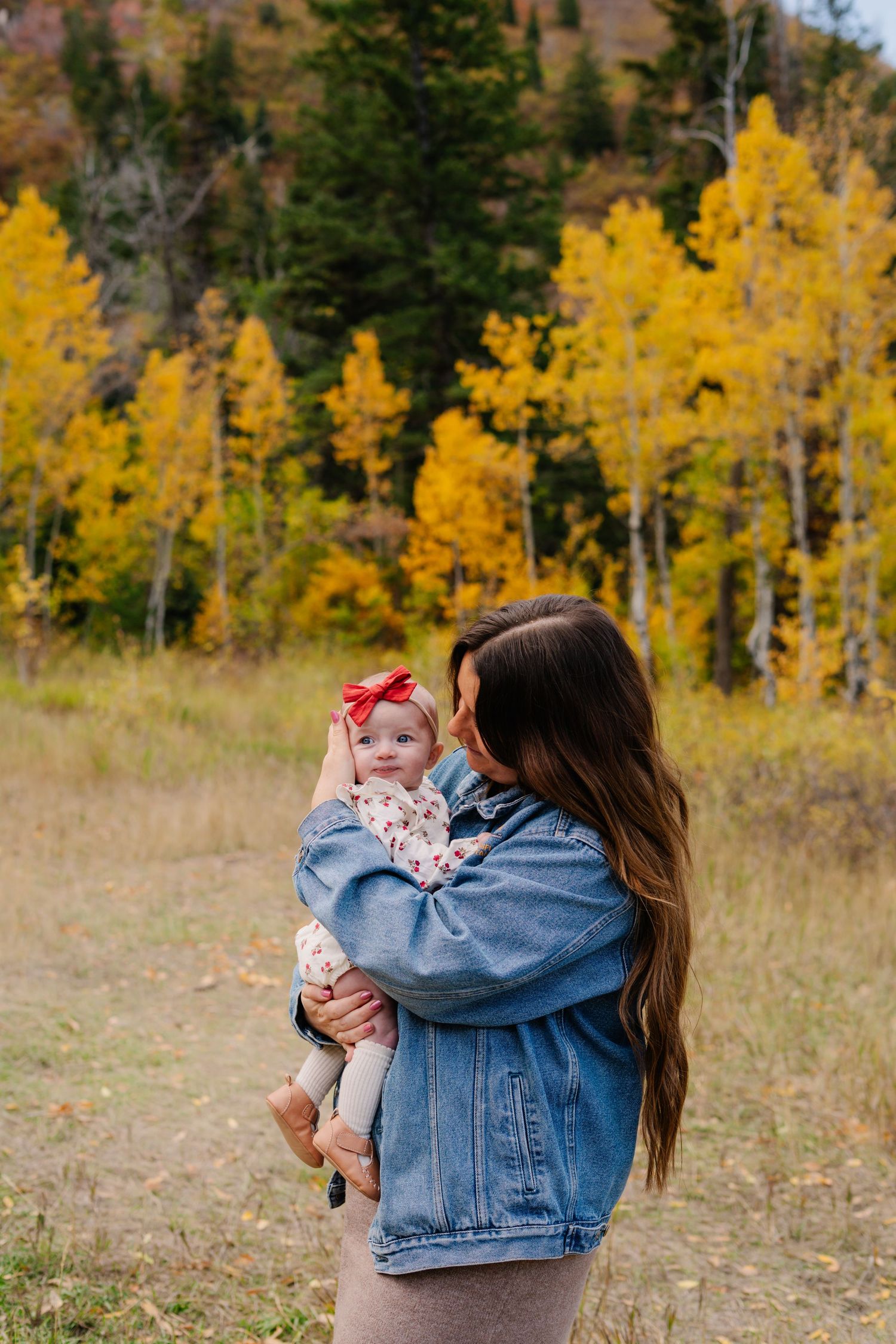 A mother in a denim jacket holds her baby girl with a red bow amid vibrant yellow autumn aspen trees.