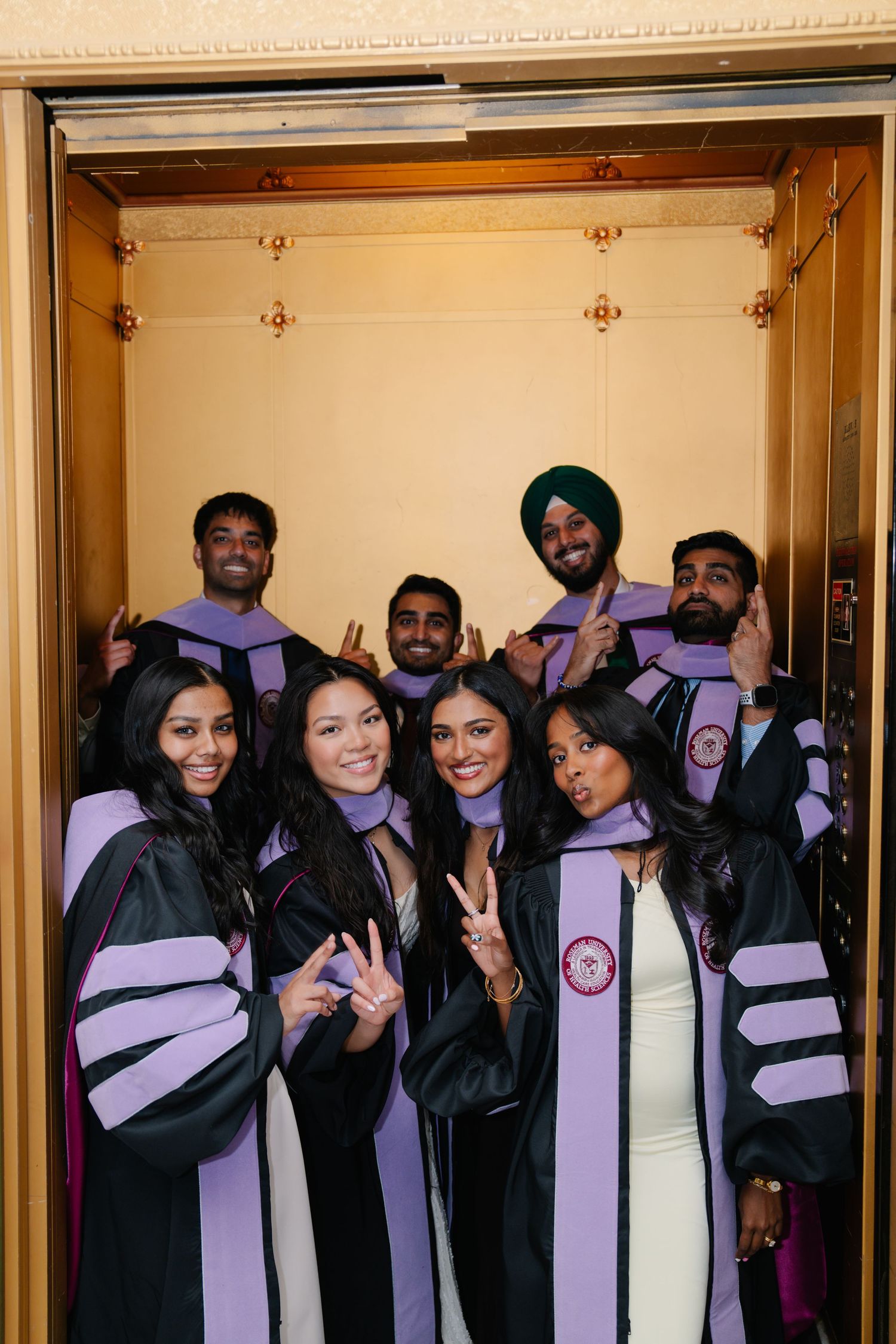 Group of graduates in black and purple academic robes posing together inside an elevator celebrating their graduation day.