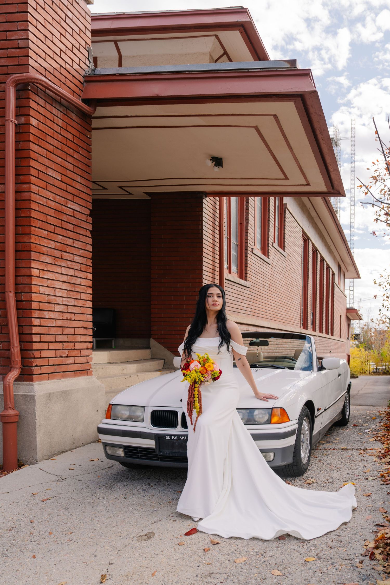 Bride in white wedding gown holding colorful bouquet, posing beside a white BMW convertible outside a brick building.