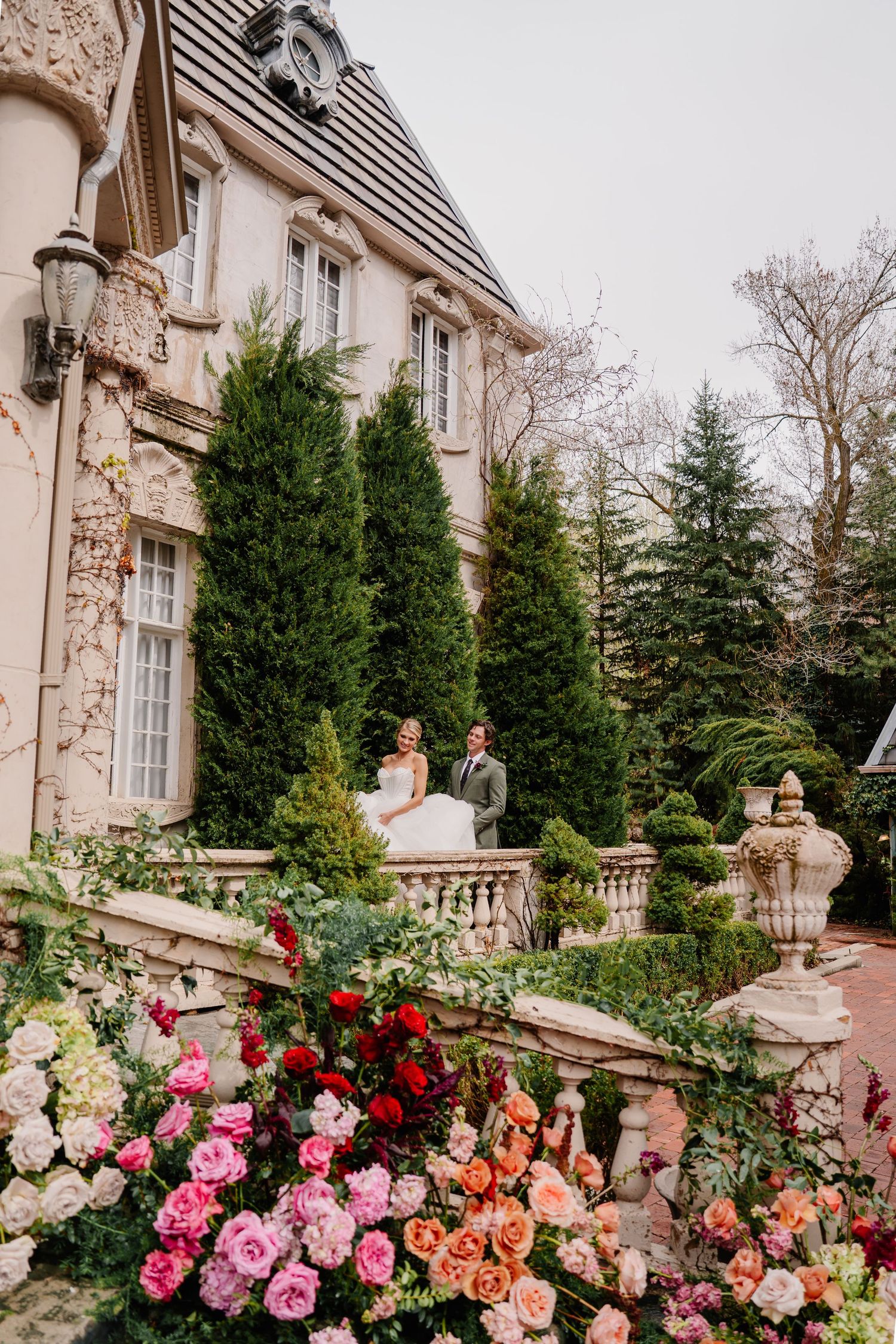 Vibrant pink and coral roses bloom along stone balustrades of a grand historic mansion.