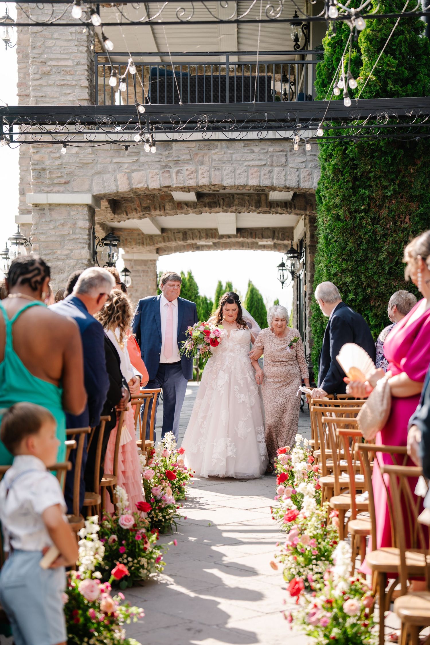 Wedding ceremony aisle lined with pink flowers and wooden chairs under a stone archway.