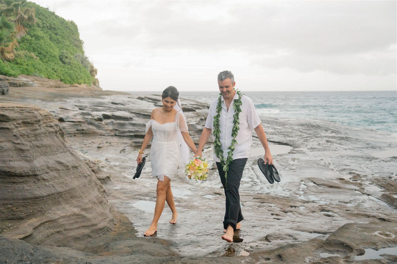 Newlywed couple walks barefoot along a rocky beach in tropical destination — captured with a storytelling, editorial style