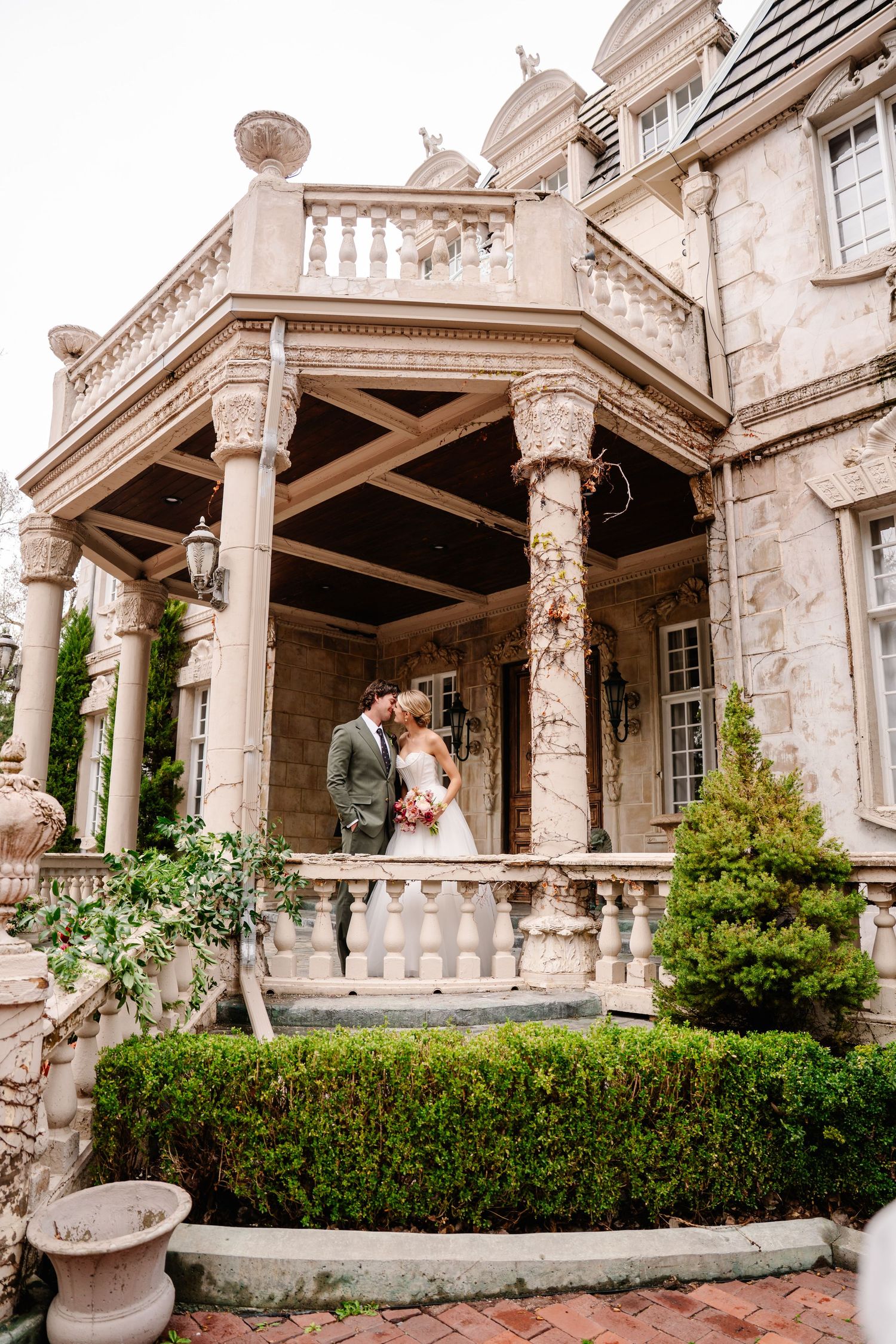 Elegant stone mansion veranda with ornate columns and manicured hedges creates a romantic wedding venue setting.