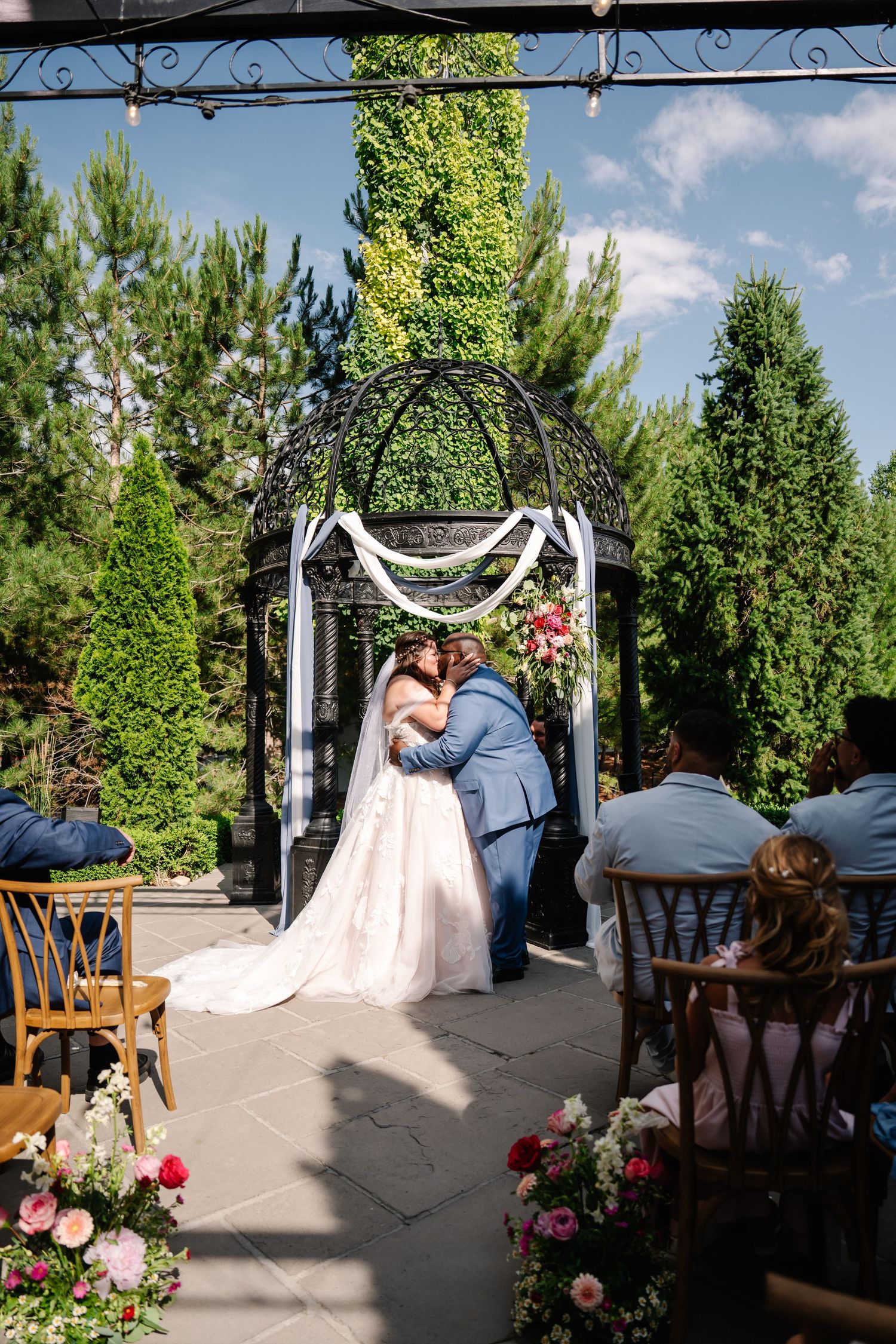 Wedding ceremony under a decorated metal arch with floral arrangements in an outdoor garden setting.