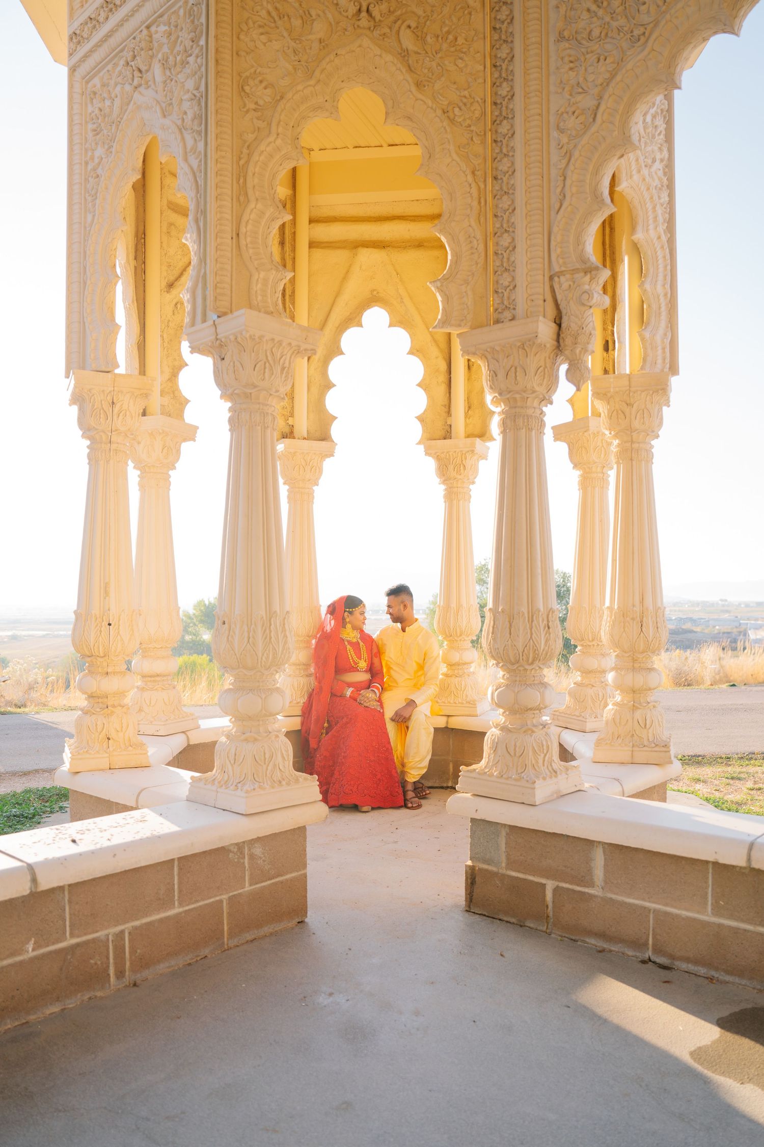 A couple in traditional attire sitting in an ornate white marble pavilion during golden hour.