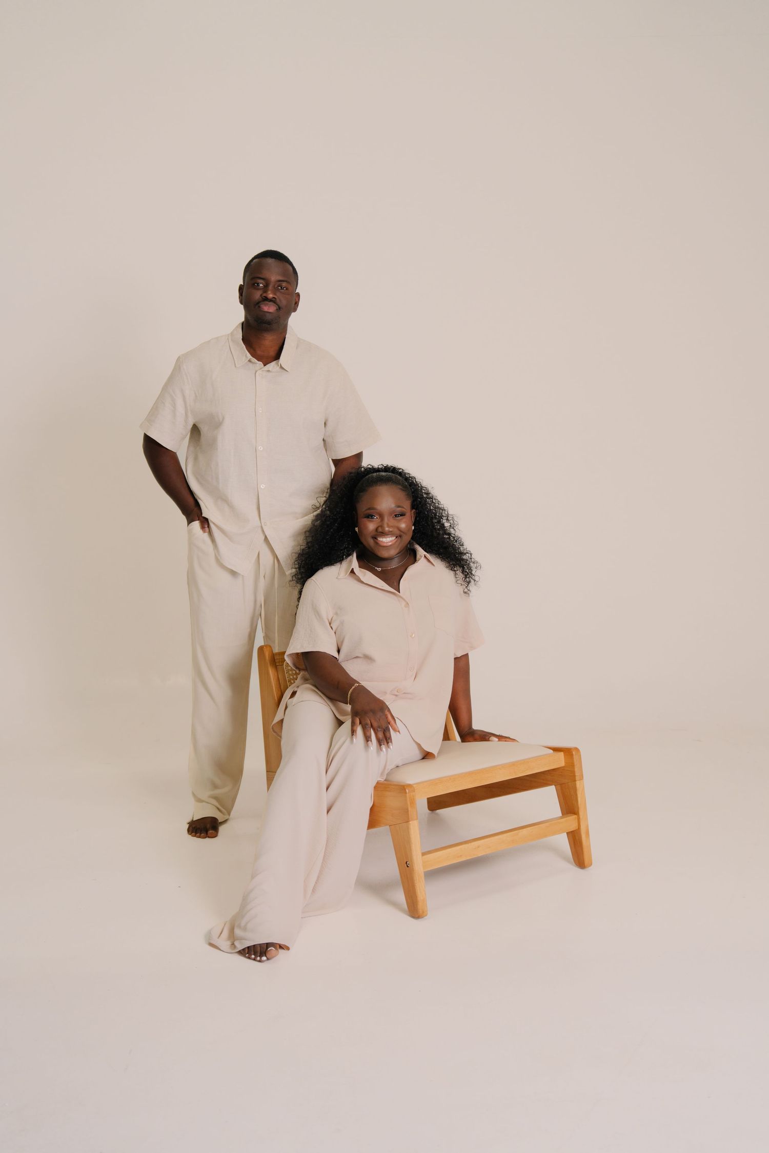 A man and woman in matching beige outfits pose together in a studio, woman seated on wooden stool, smiling.