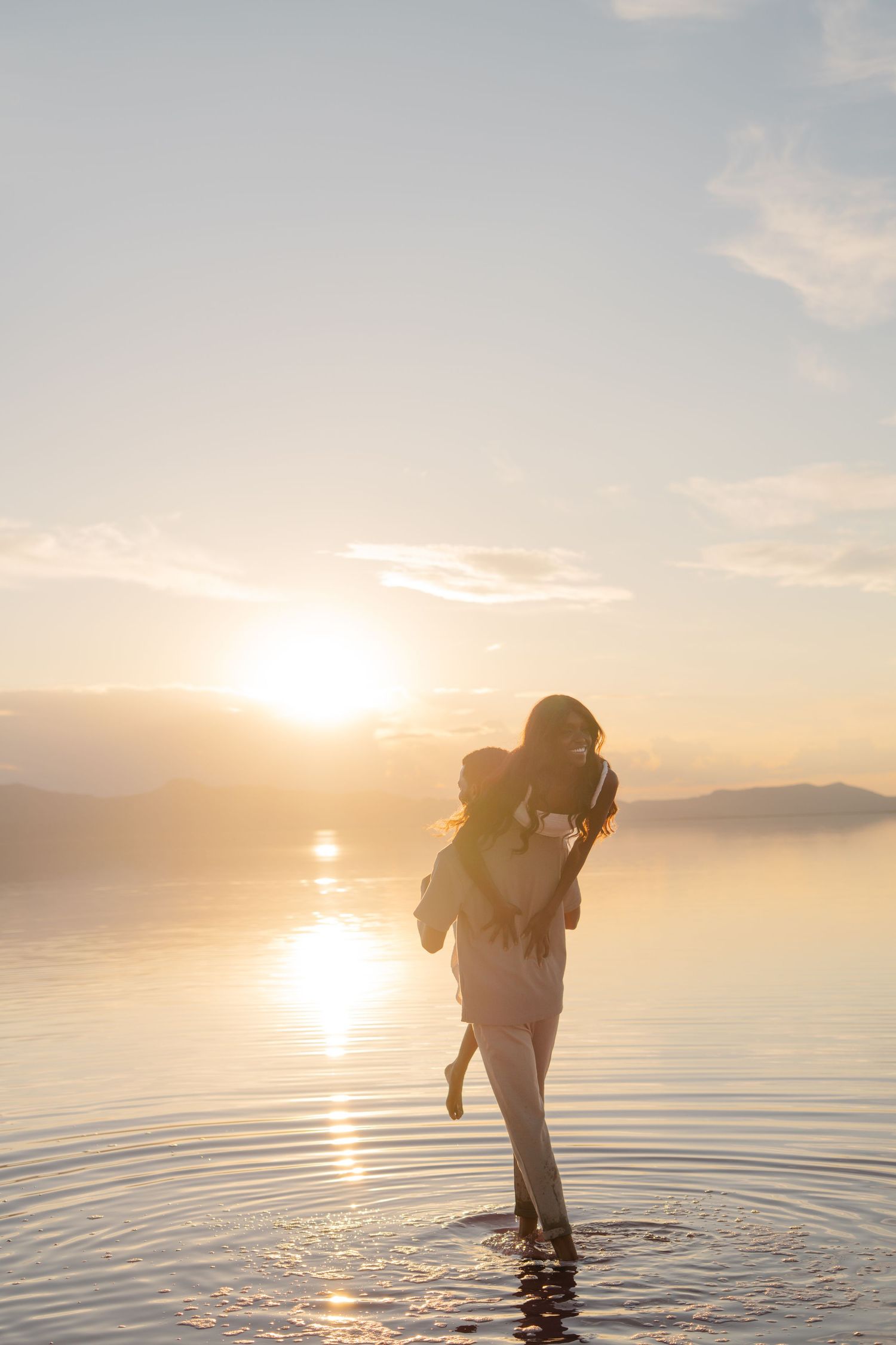 A mother carries her child while standing in shallow water during a golden sunset over calm lake waters.