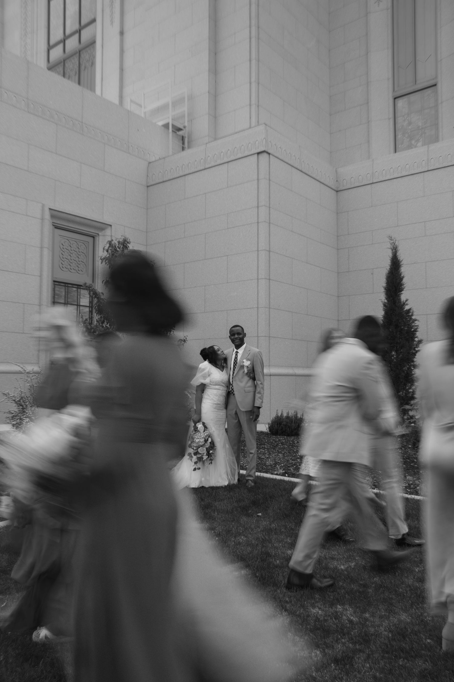 Black and white wedding photo of a couple posing outside a building while guests walk by in motion blur.