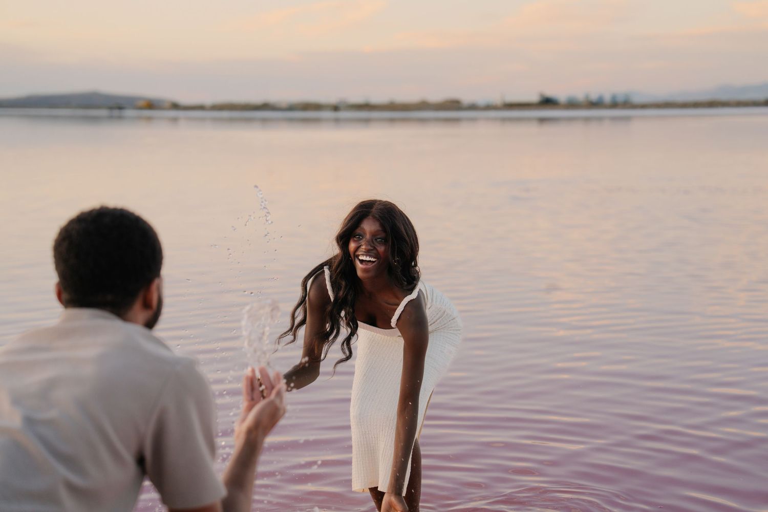A couple shares a joyful moment while wading in pink-tinted water at sunset.