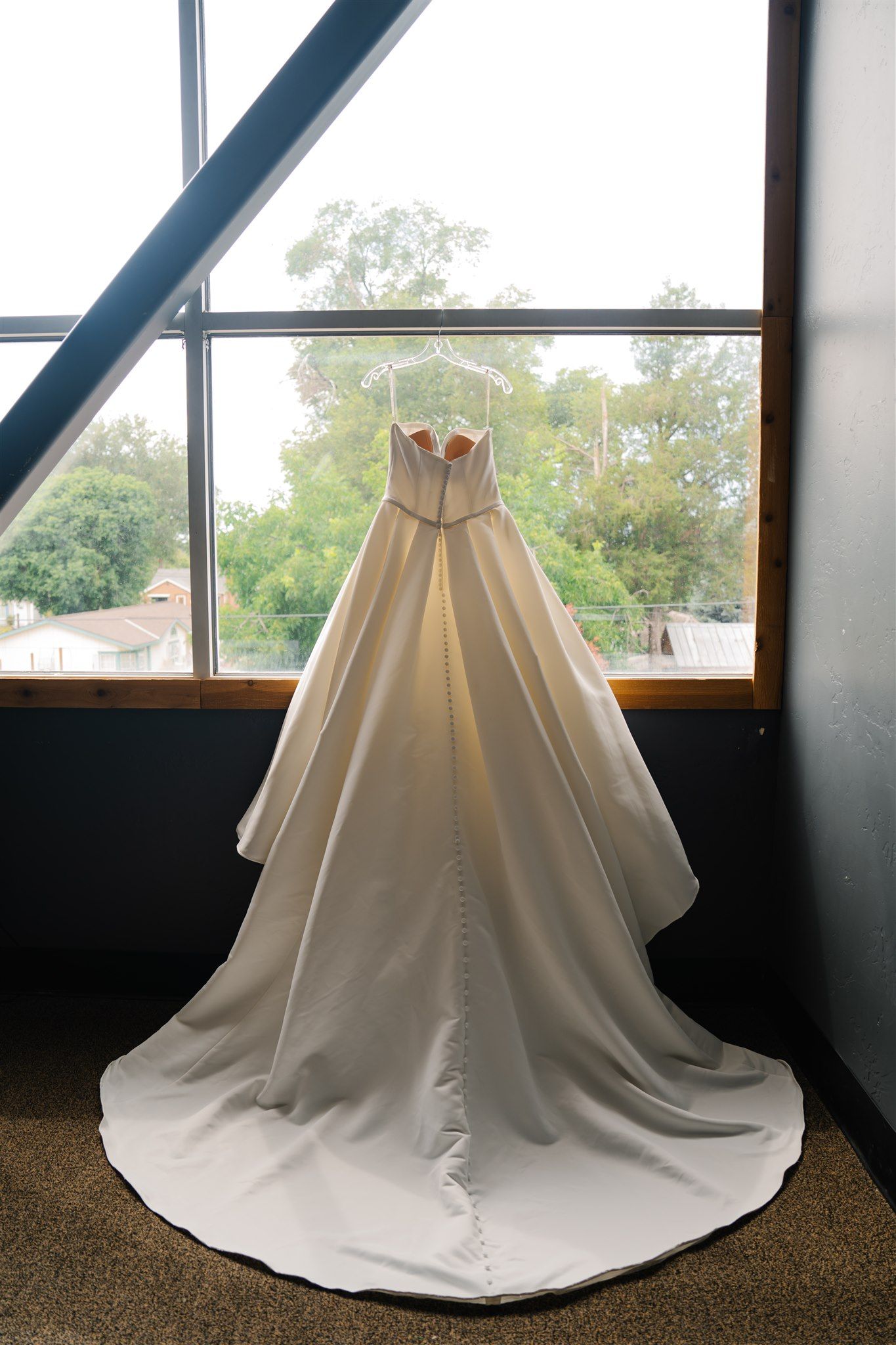 A white wedding dress train spread out on carpeted floor near windows.