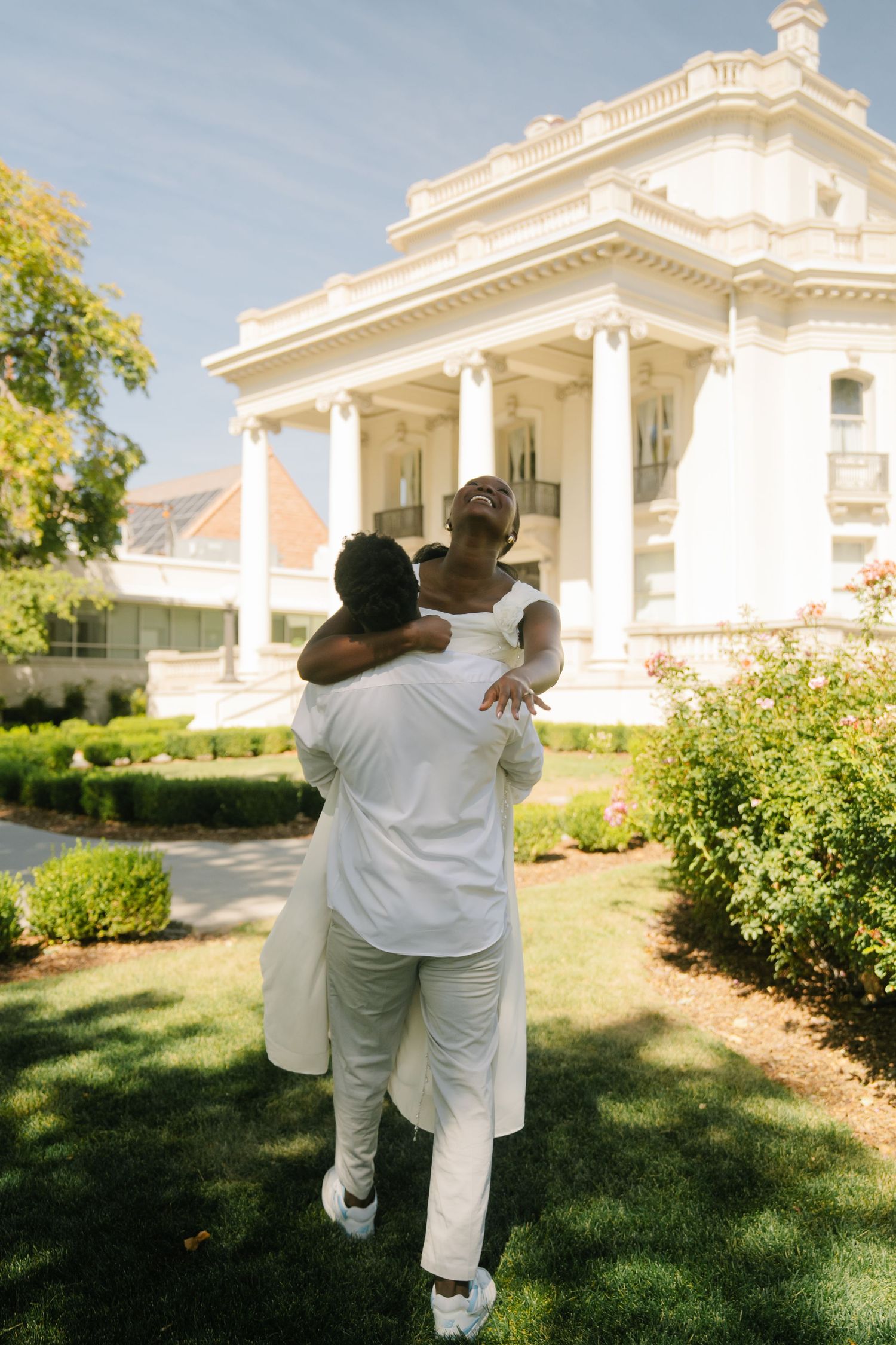 Couple in white outfits embracing joyfully in front of an elegant white mansion with columns on a sunny day.