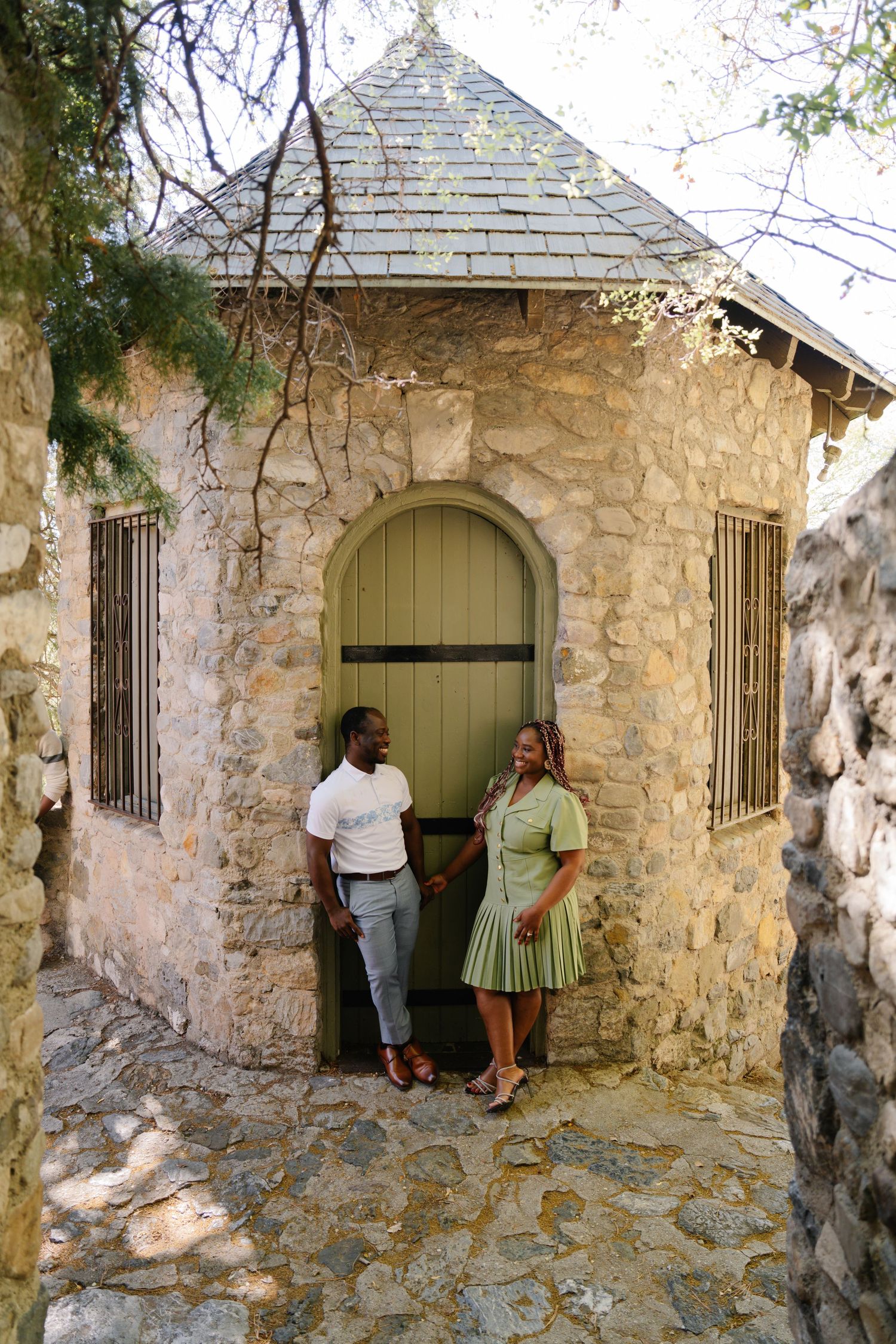 Couple standing together in front of a rustic stone building with a green arched door on a cobblestone path.