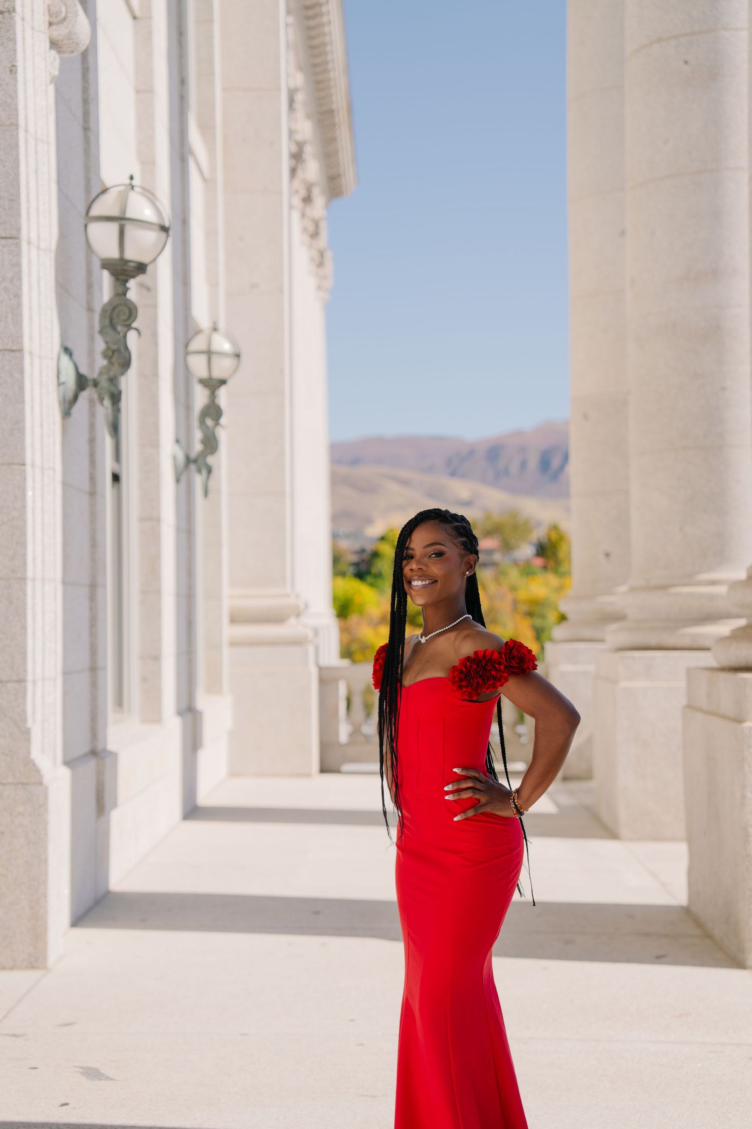 Young woman in elegant red off-shoulder gown poses confidently between white columns with mountain backdrop.