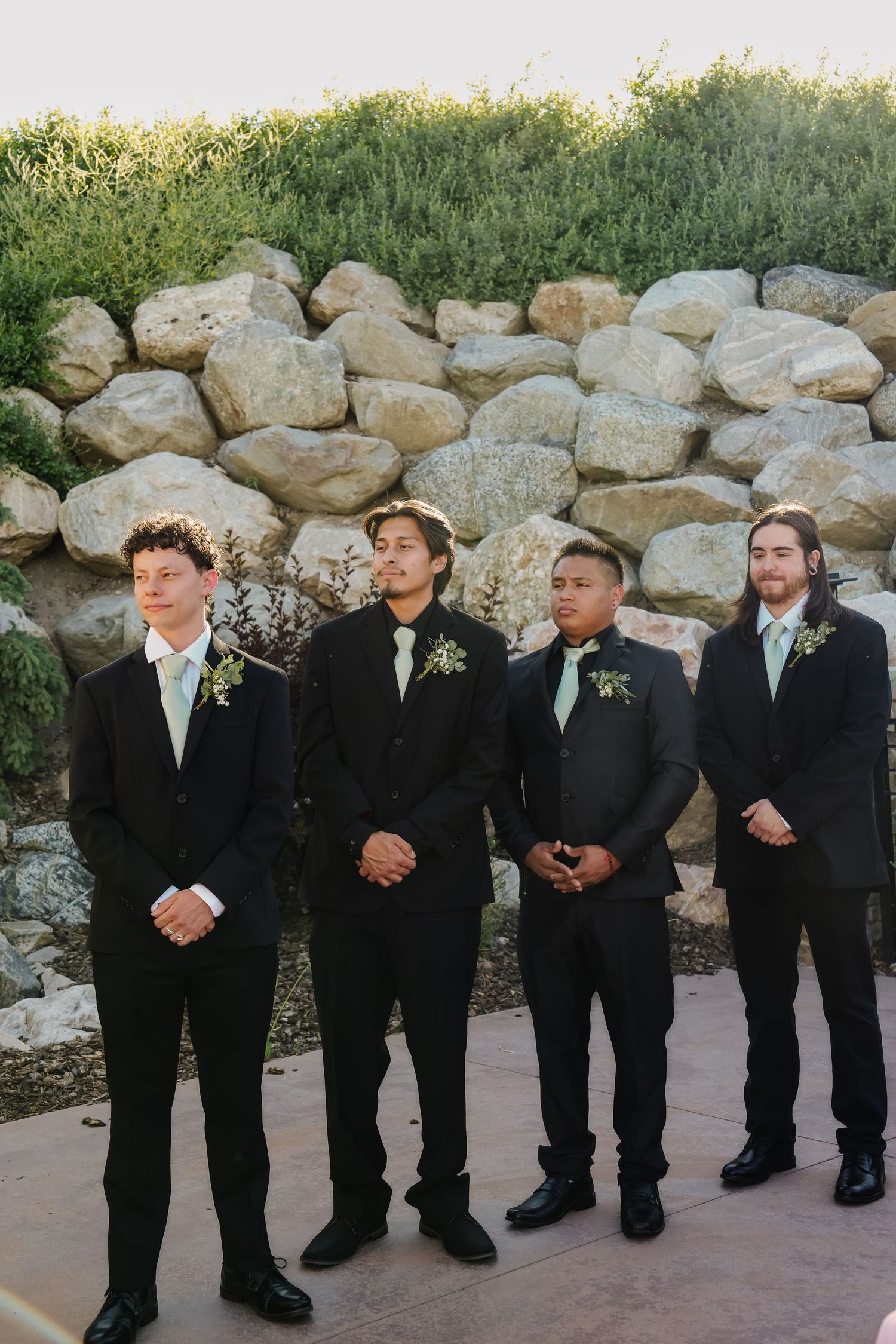 Groomsmen in black suits and ties lined up against stone wall.
