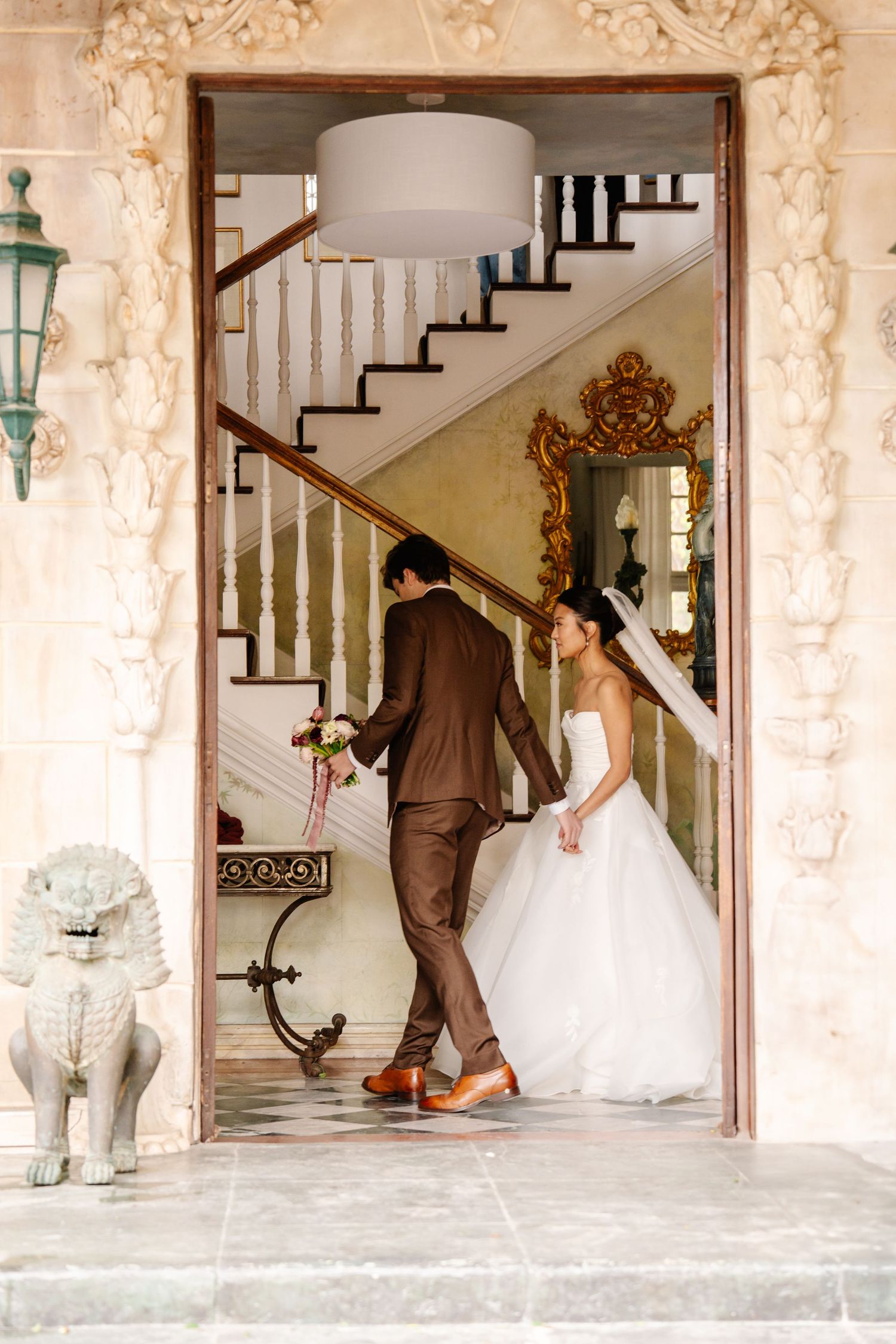 A couple in wedding attire playfully ascends an ornate staircase with vintage decor.