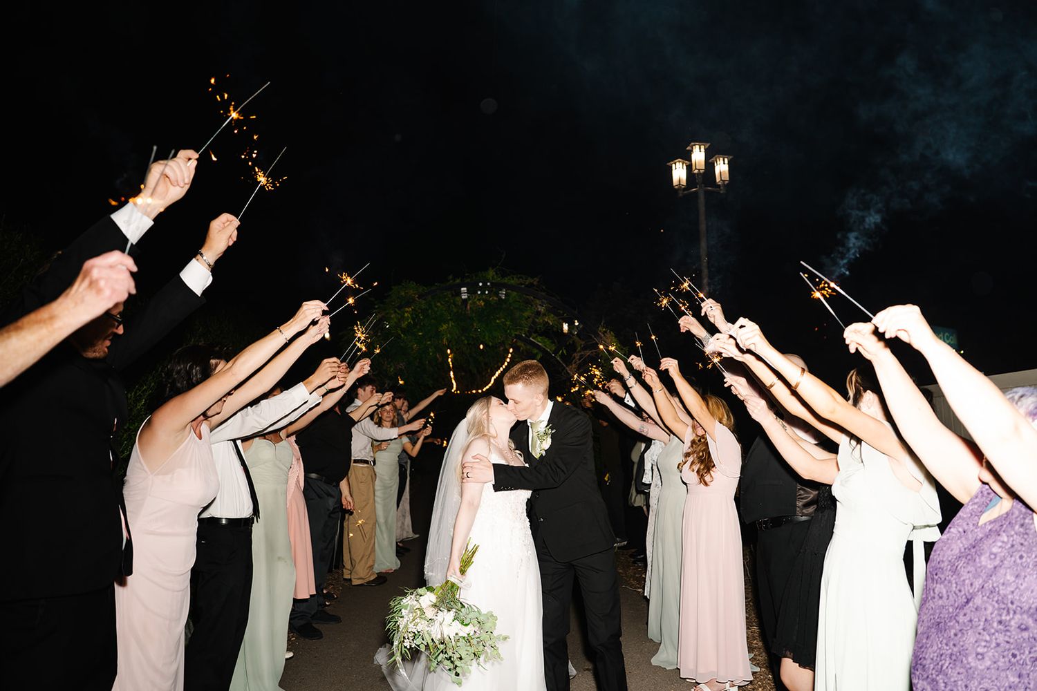 Wedding guests raise sparklers in celebration as newlyweds walk through a sparkler exit at nighttime.