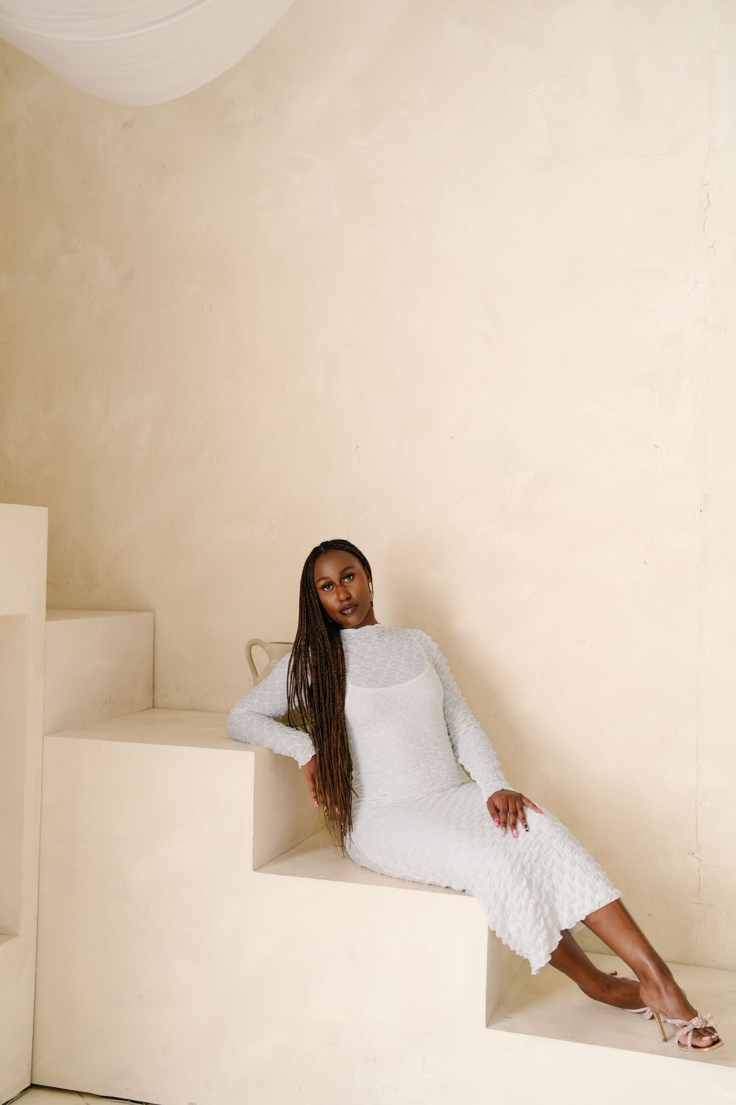 A Black woman with long braids sits gracefully on white steps wearing an elegant white textured midi dress with heels.