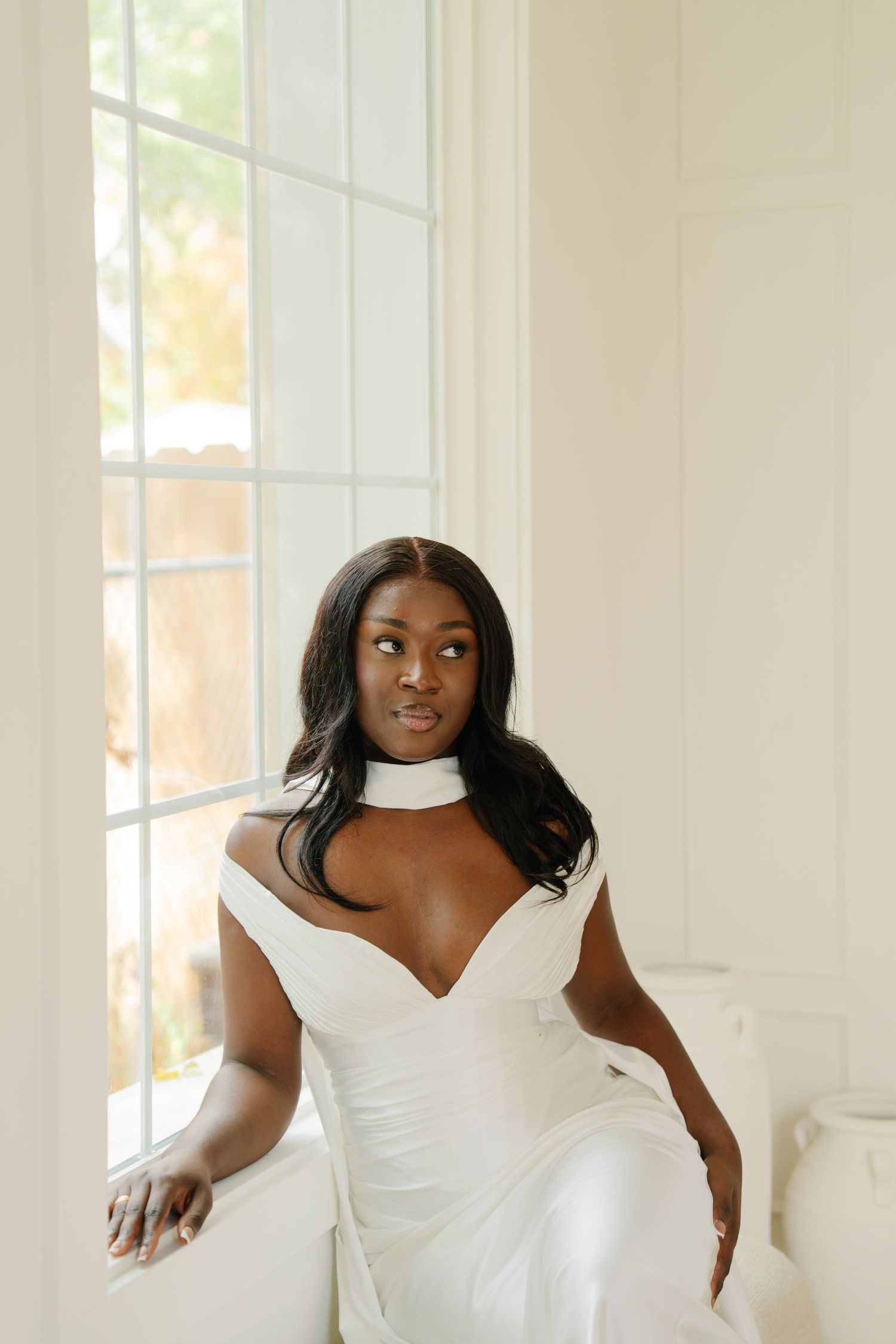 A Black bride in a white off-shoulder gown with choker neckline poses elegantly by a bright window.