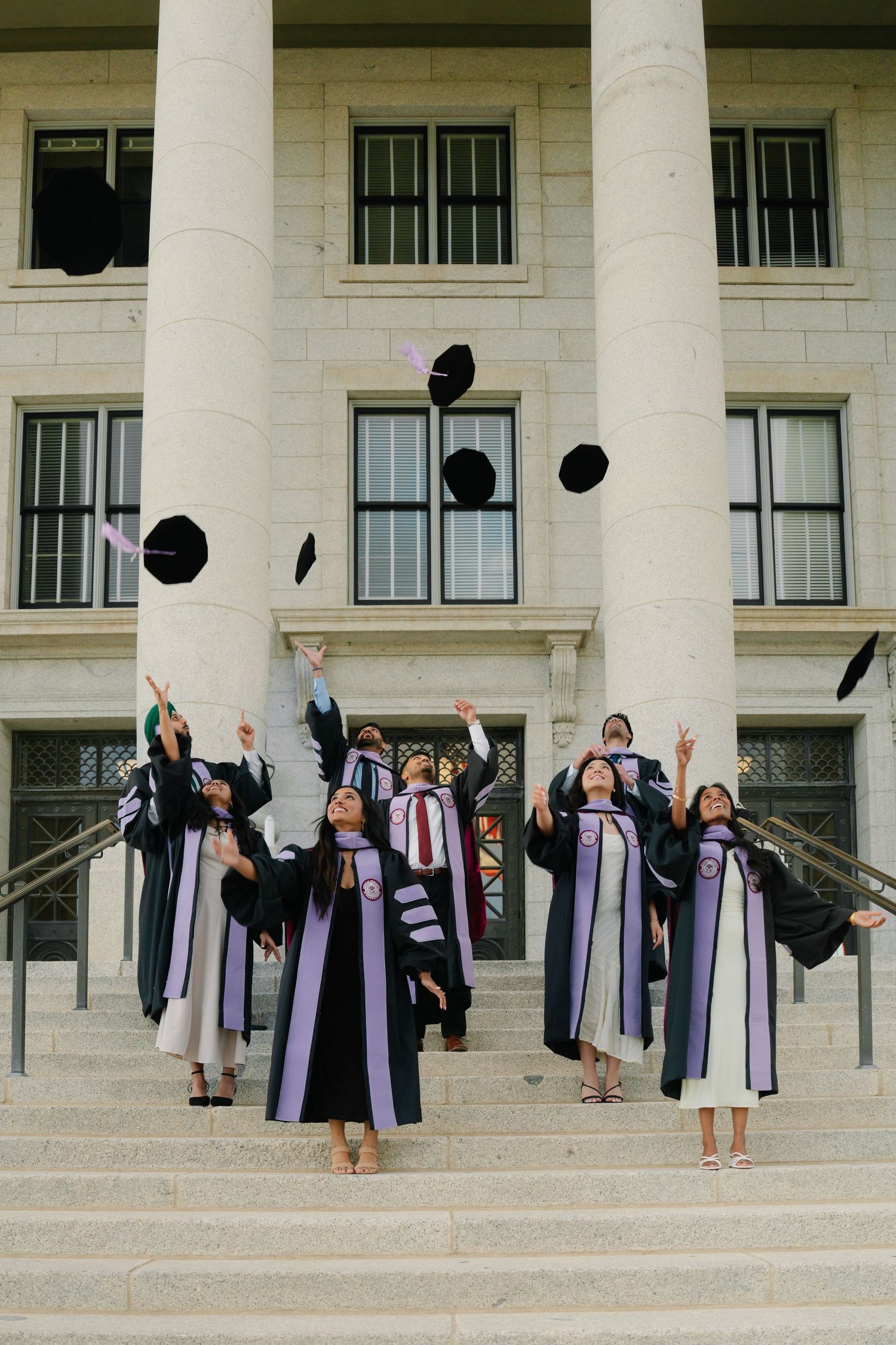 Graduates in black and purple robes toss caps in the air on steps of a classical building during graduation celebration.