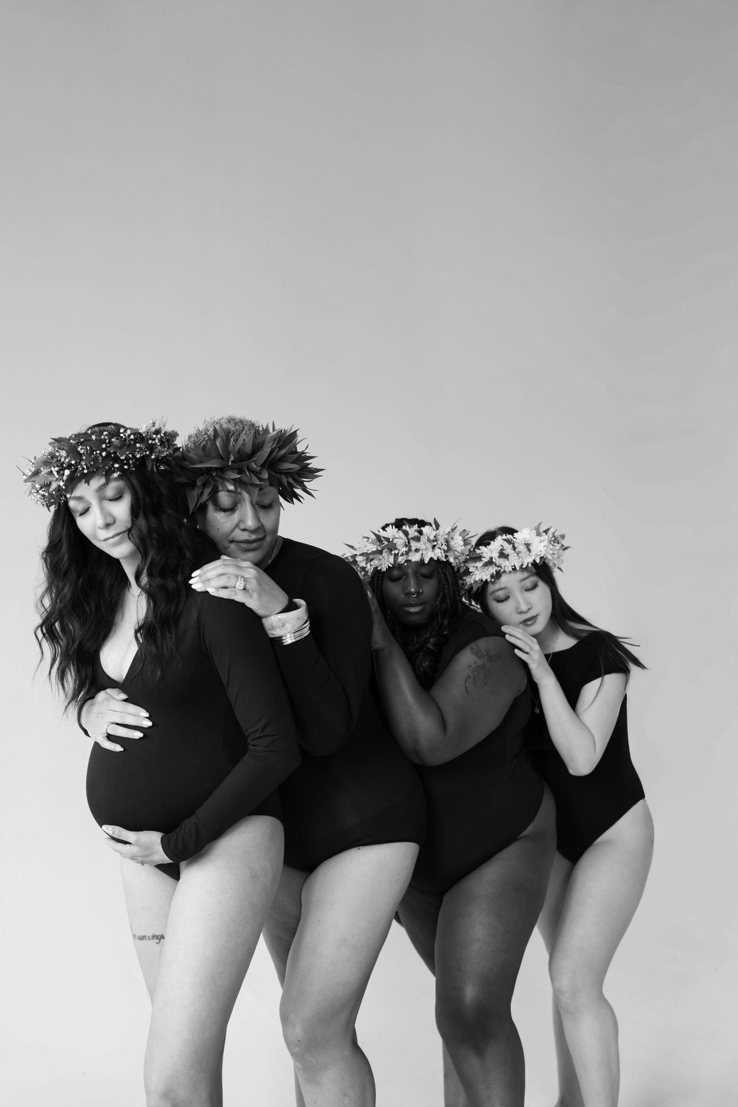 Four diverse women in black bodysuits and floral crowns pose together, one visibly pregnant, in a black and white studio photo.