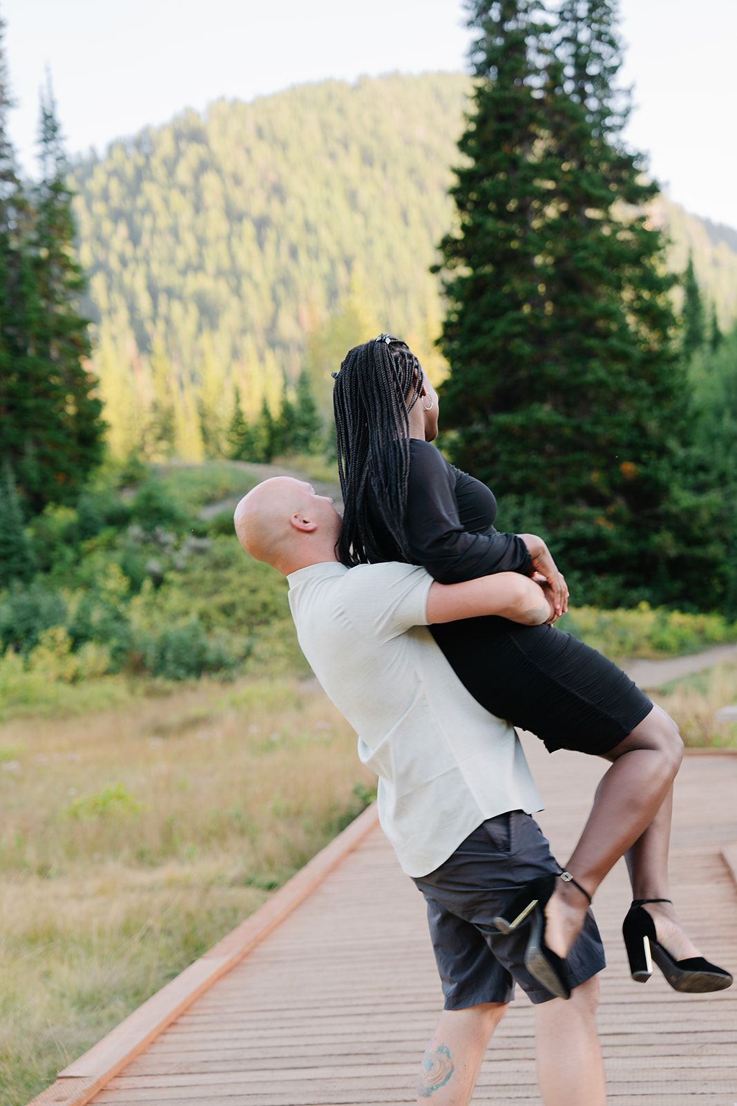 A playful couple dances together on a mountain boardwalk surrounded by evergreen trees.