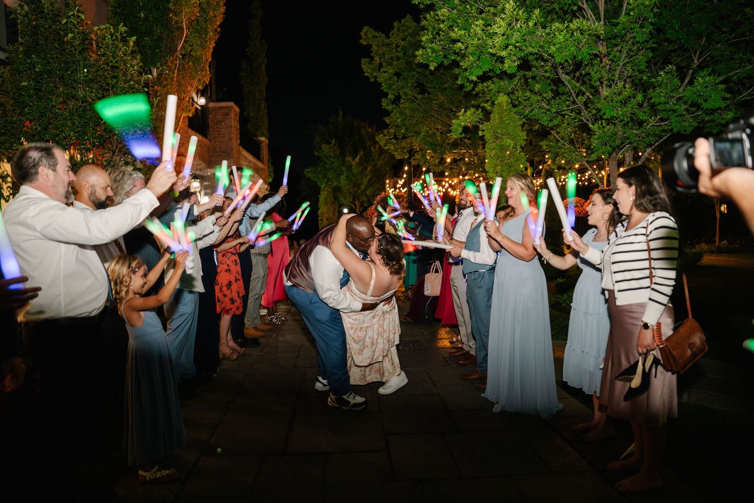 Nighttime wedding celebration with guests dancing and waving glow sticks outdoors.