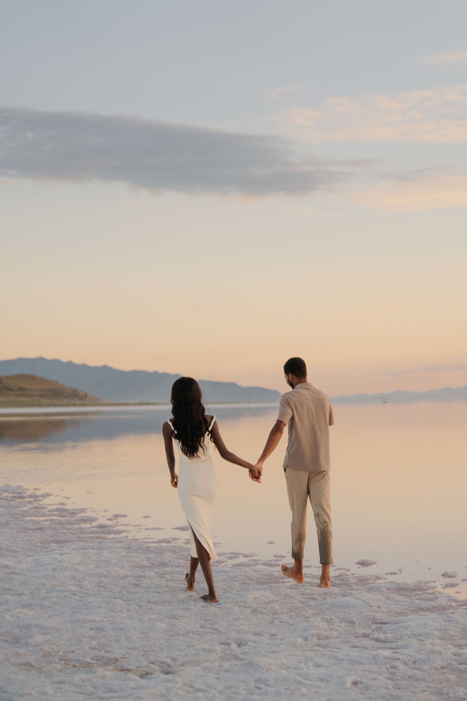 Two people walking hand in hand through shallow pink waters at dusk.