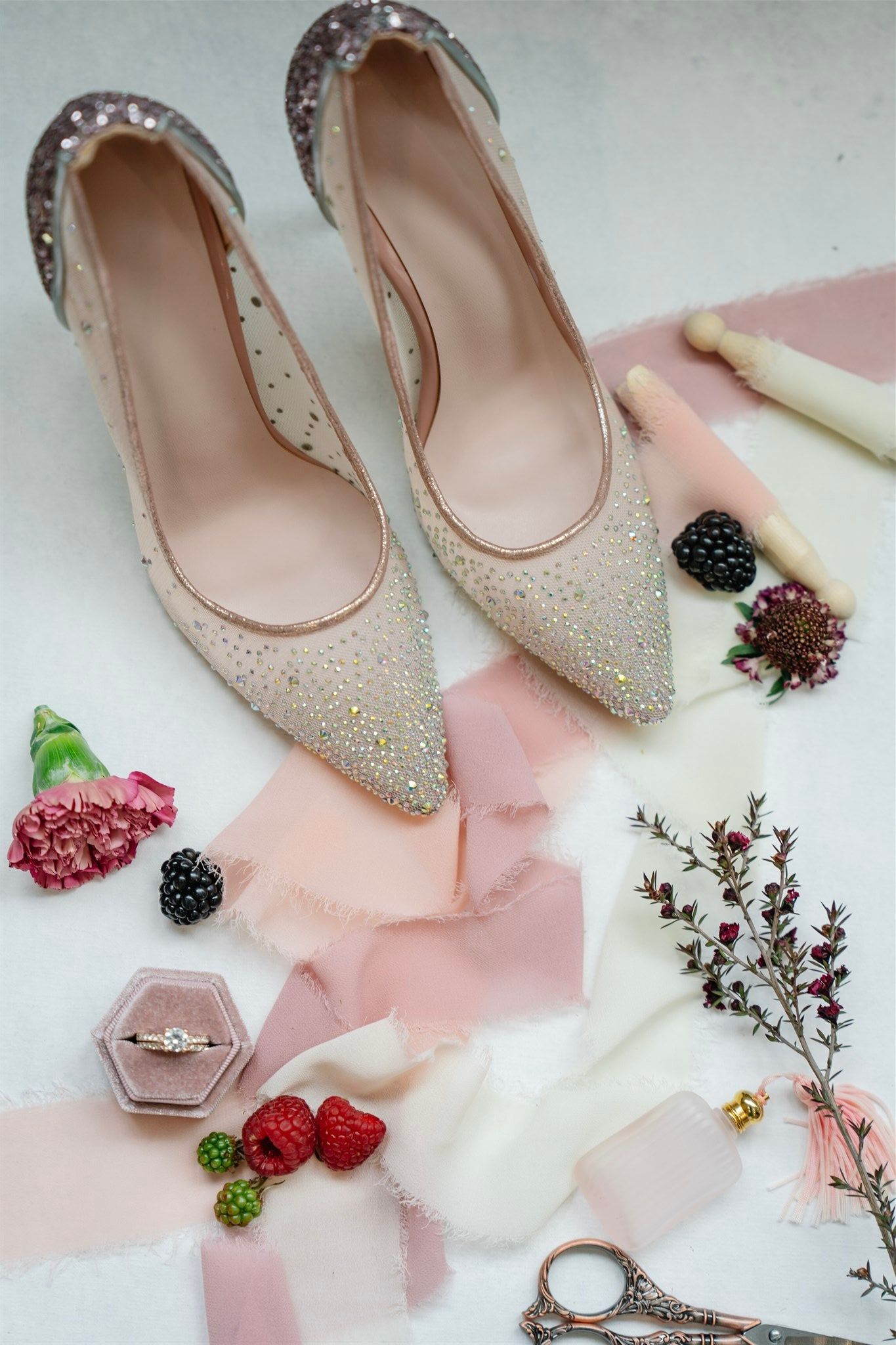 Sparkly bridal shoes with engagement ring and berries styled on pink and white fabric.