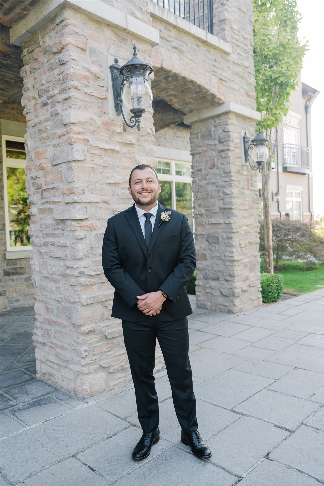 A groomsman in a black suit poses in front of a stone building facade.