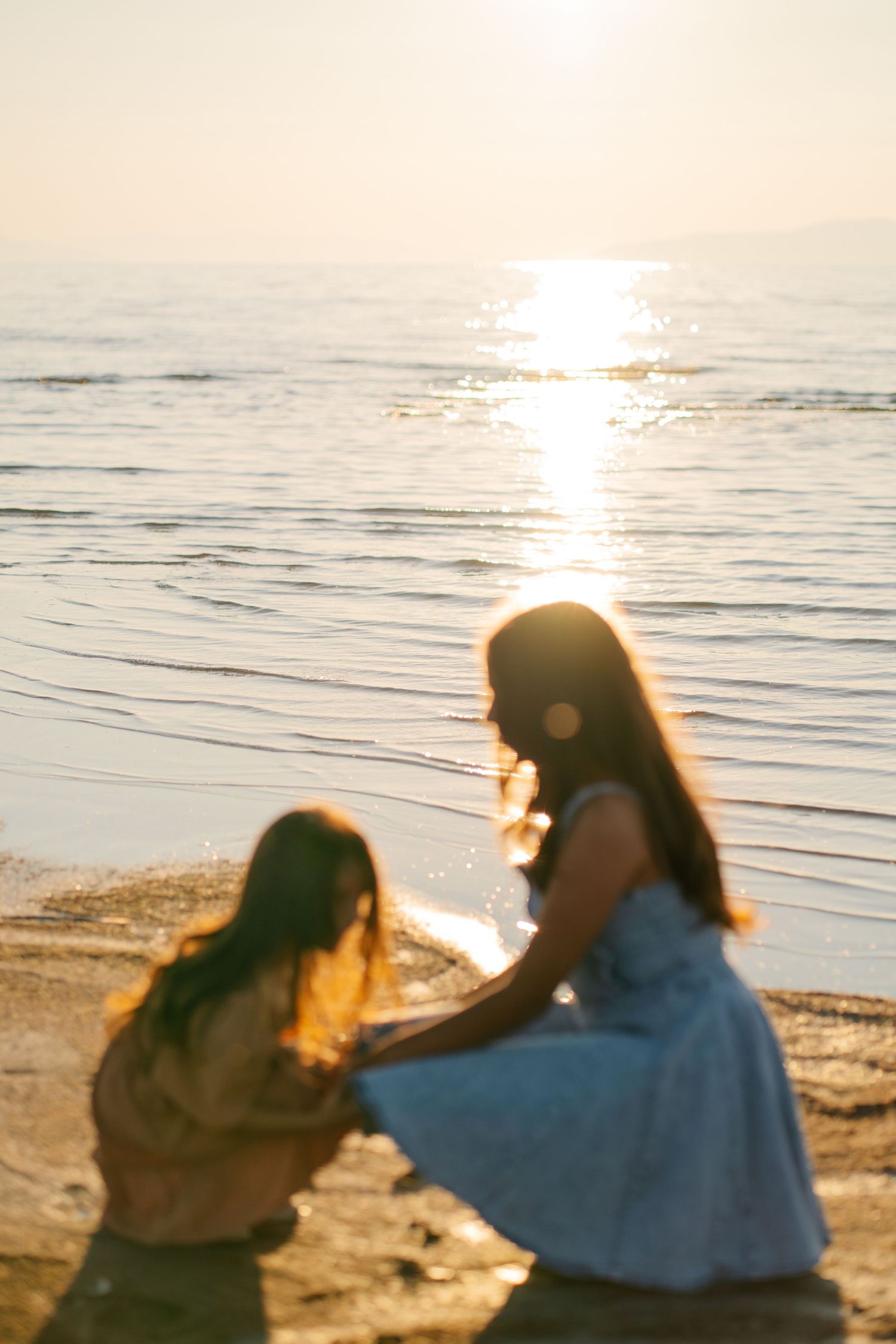 Two women sitting on a sandy beach at golden sunset, with calm ocean waves reflecting warm sunlight behind them.