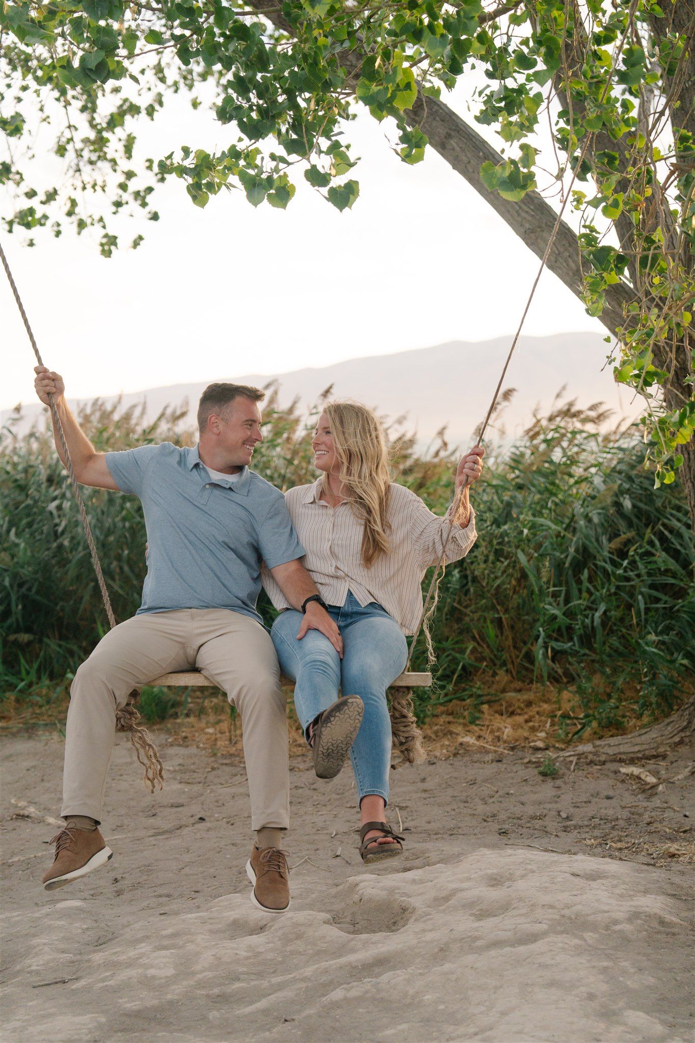 A playful couple swings together on a wooden swing at the beach during sunset.
