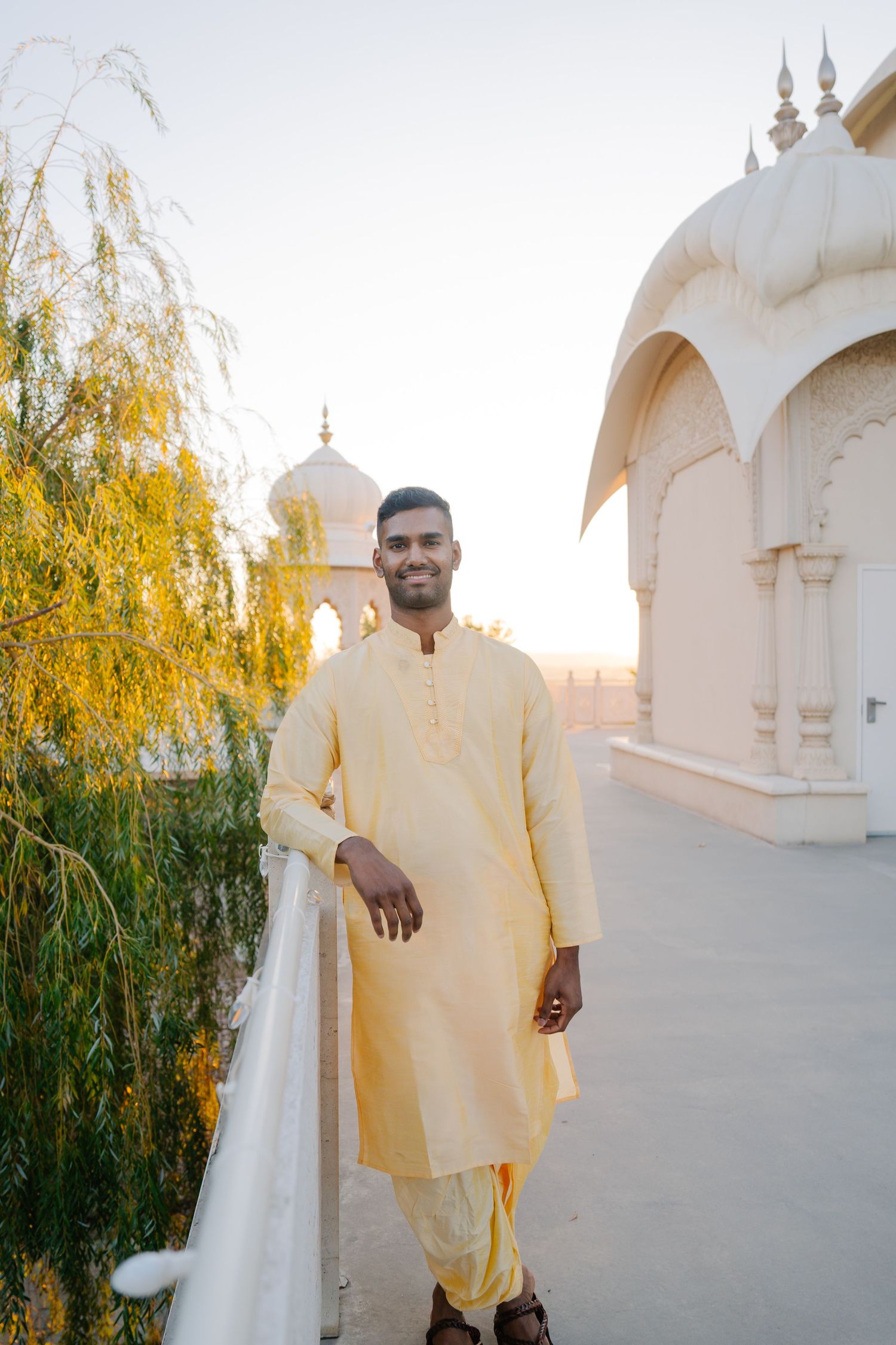 A person in a cream kurta stands beside a temple with ornate white domes and yellow willow trees.