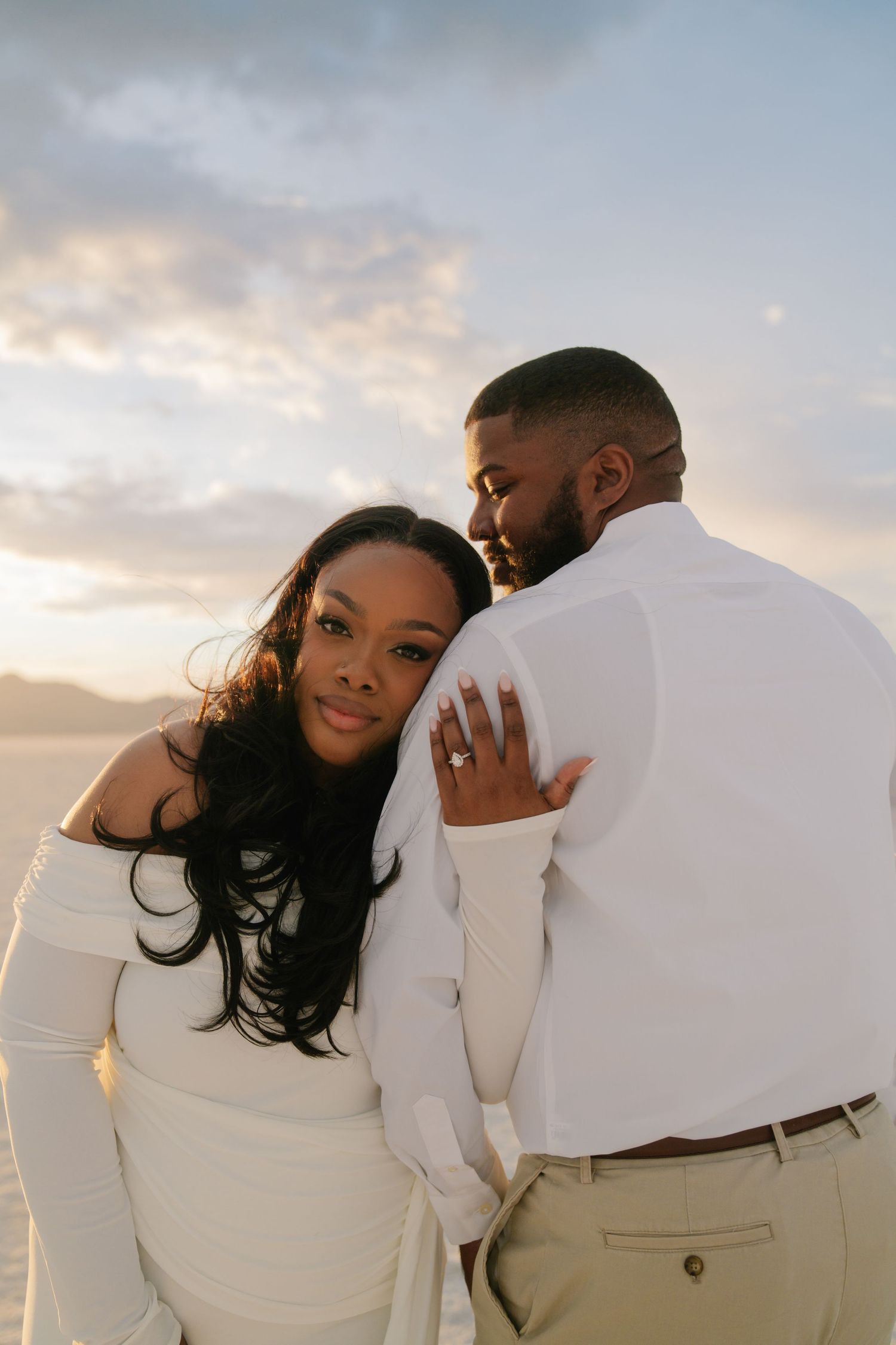 A couple in white outfits embraces at sunset, the woman showing her engagement ring while leaning against her partner.