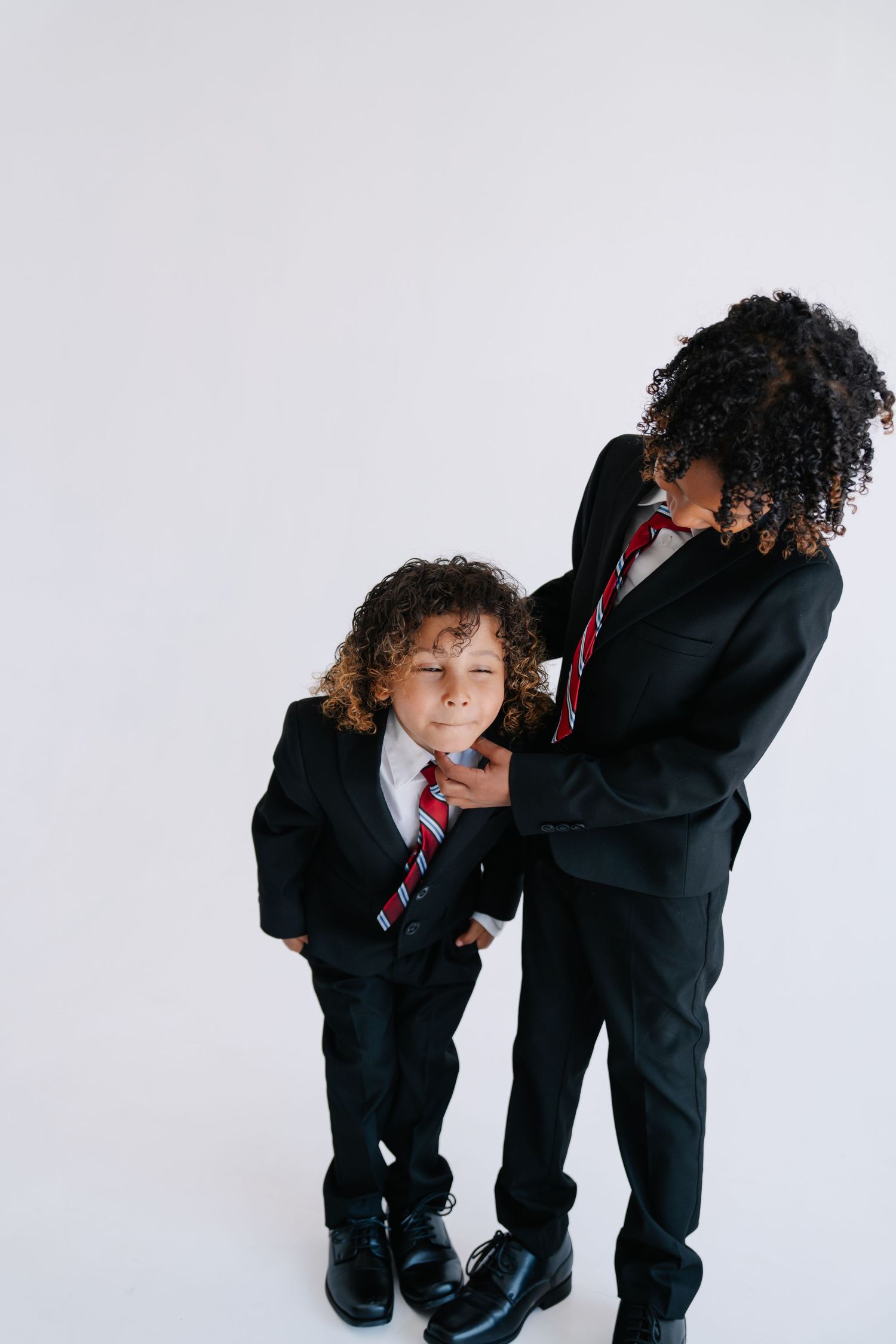 Two curly-haired children in matching black suits and striped ties pose together against a white background.