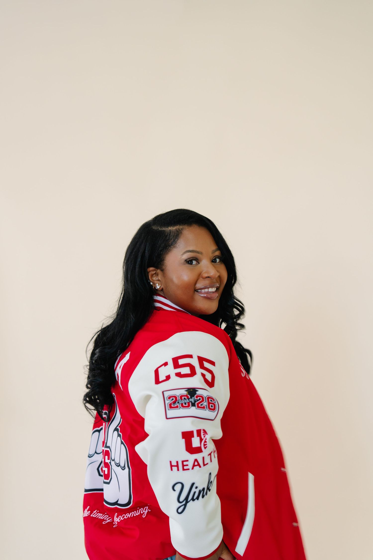 Young woman smiling over shoulder wearing red and white varsity jacket with CSS patches and U Health logo.