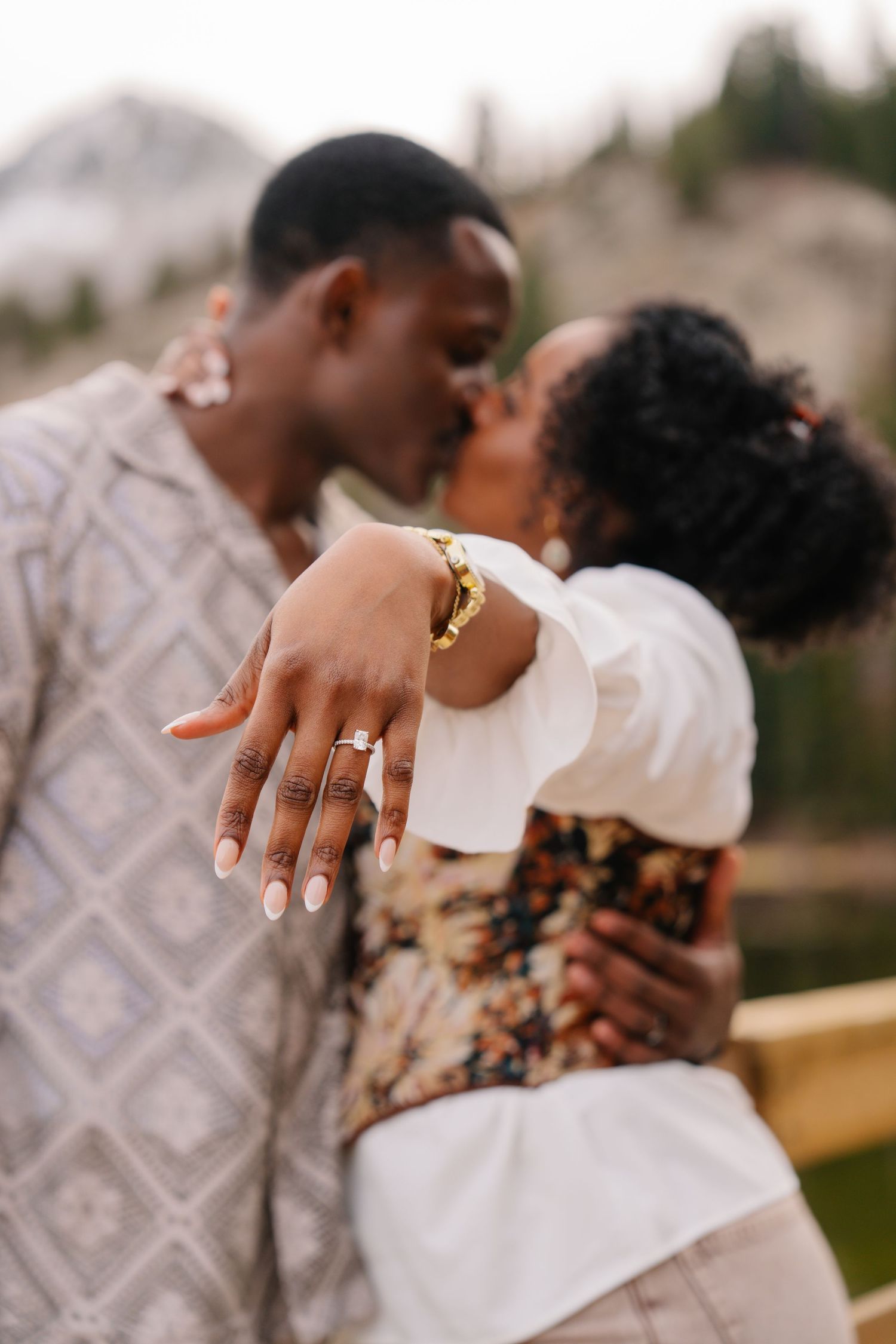 A couple shares a kiss while showing off an engagement ring, with a mountain backdrop.